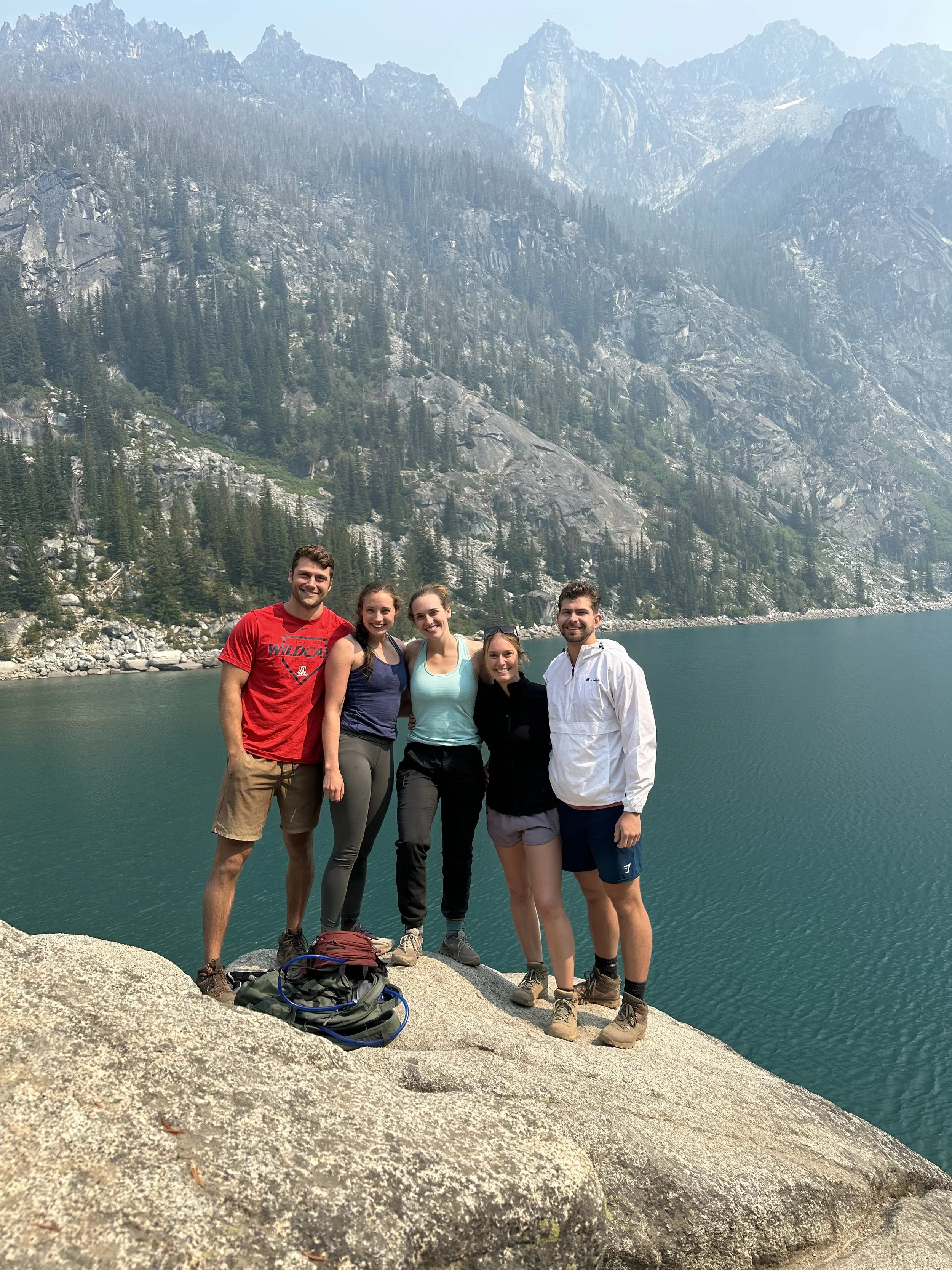 Group of five young adults standing on a large rock by a lake with forested mountains in the background, enjoying an outdoor adventure.