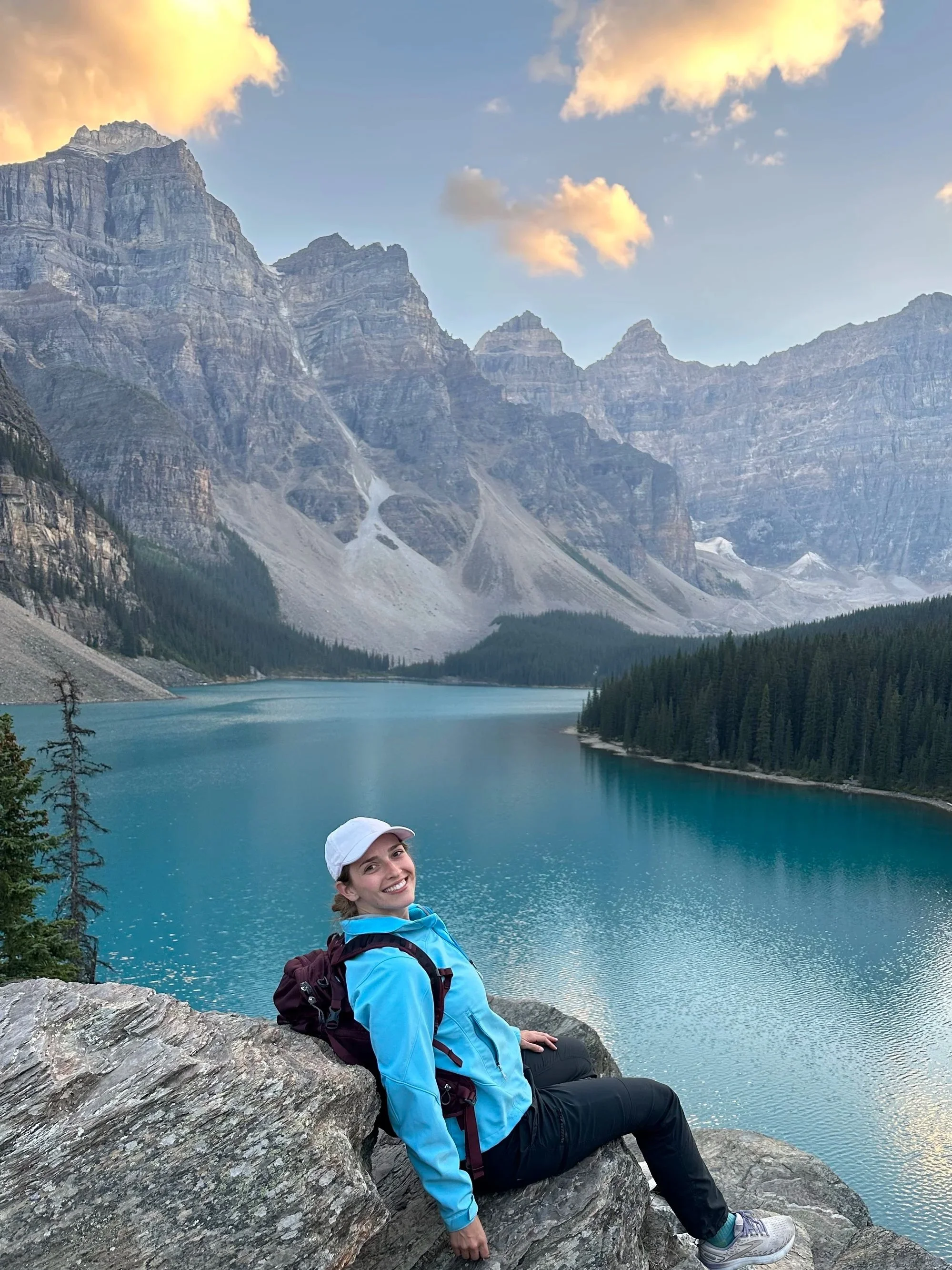 A woman smiling at the camera, sitting on a rock by a mountain lake with in the background tall peaks, trees, and a partly cloudy sky.