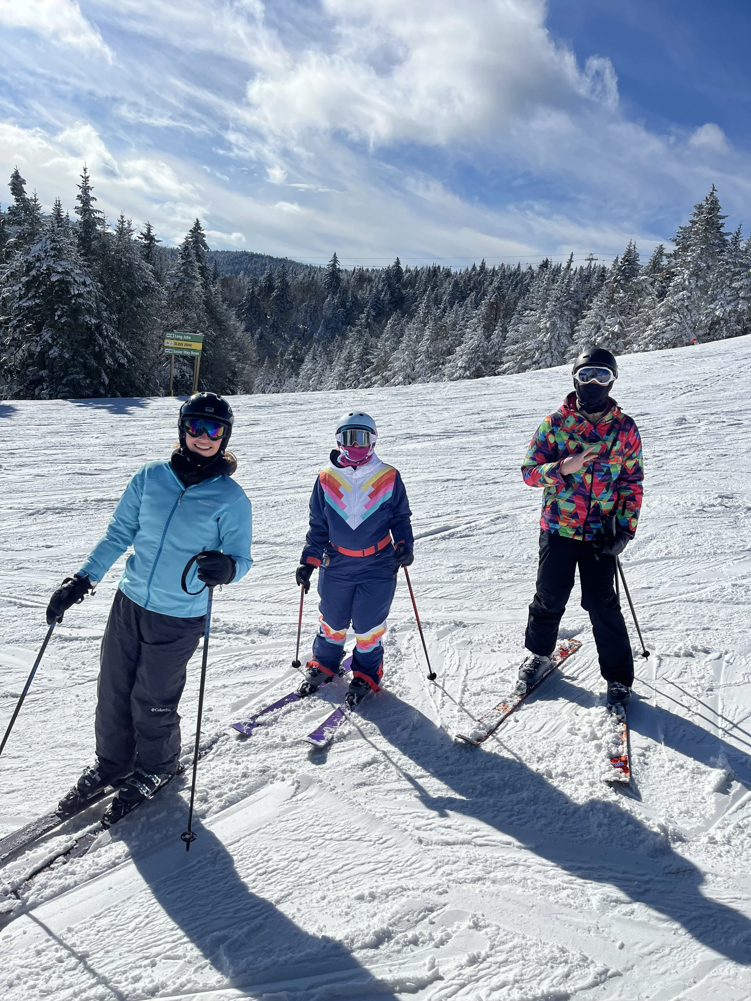 Three skiers in colorful winter gear standing on snowy ski slope with pine trees and mountains in the background under partly cloudy sky.