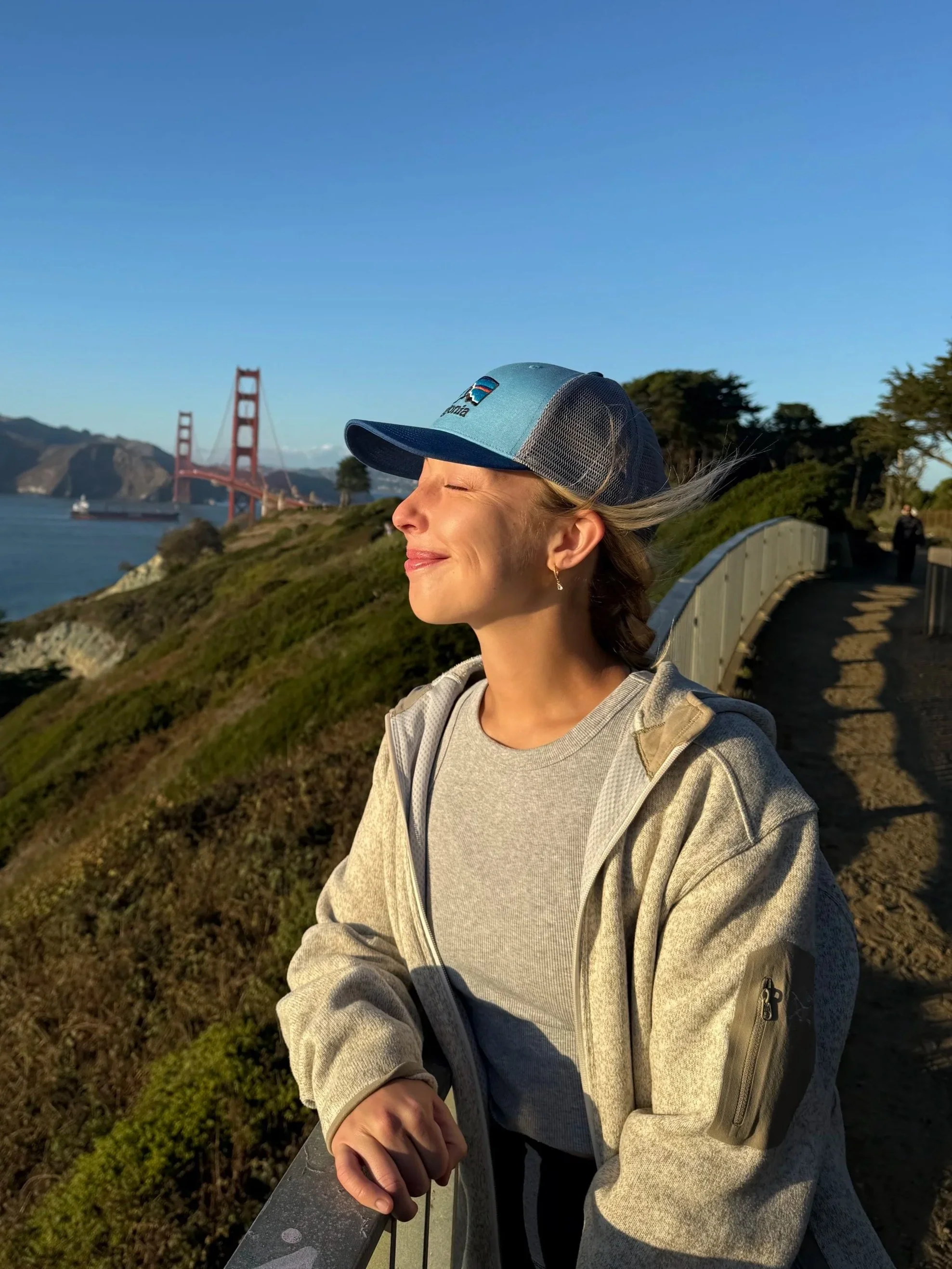 A woman with closed eyes smiling, wearing a blue cap with a Patagonia logo, standing on a scenic lookout near the Golden Gate Bridge in San Francisco during sunset.