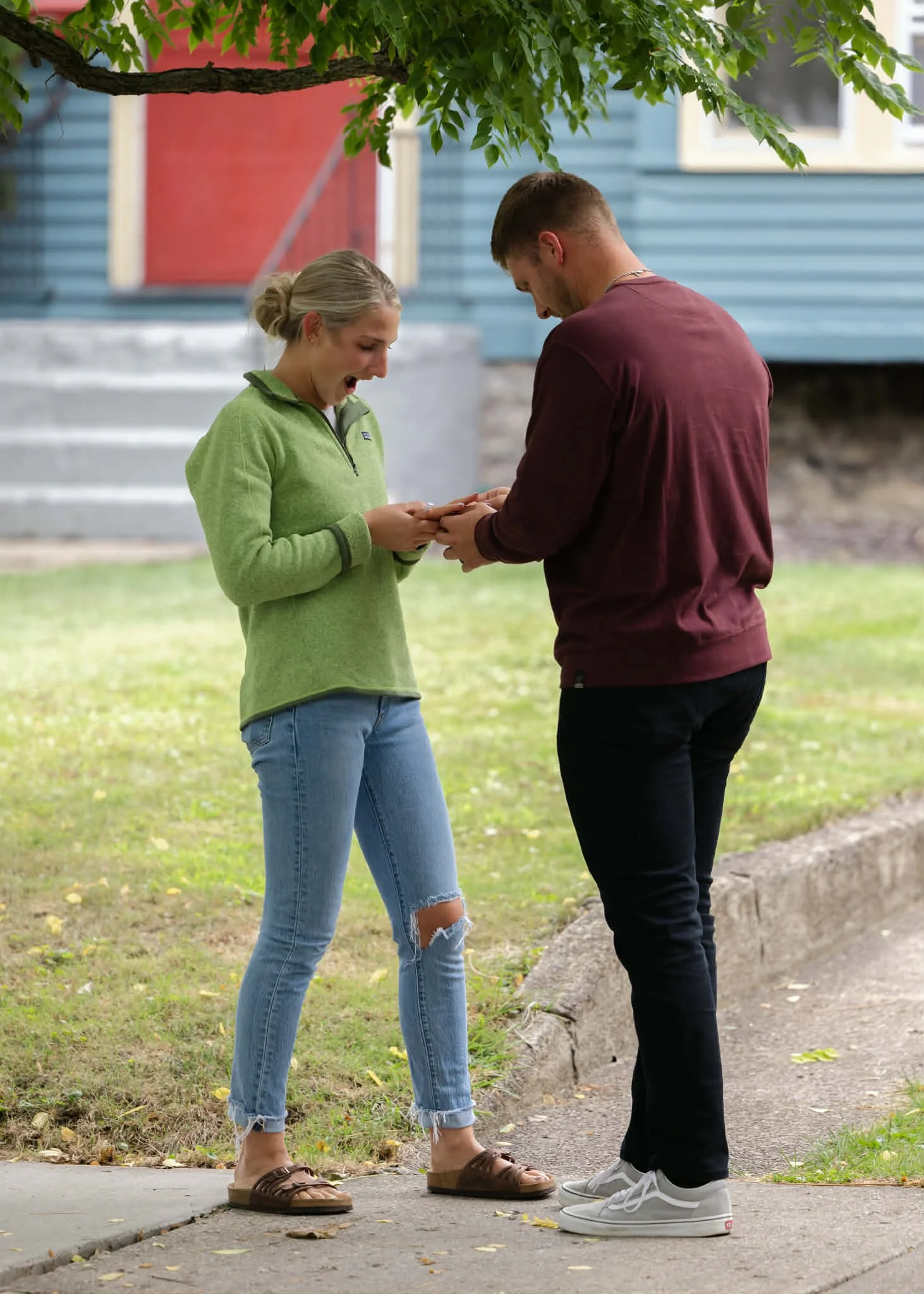 A young woman with blond hair in a bun wearing a green jacket and ripped jeans is smiling and looking at her phone, while a young man in a burgundy shirt and black pants stands next to her, also looking at her phone. They are outdoors under a tree, n