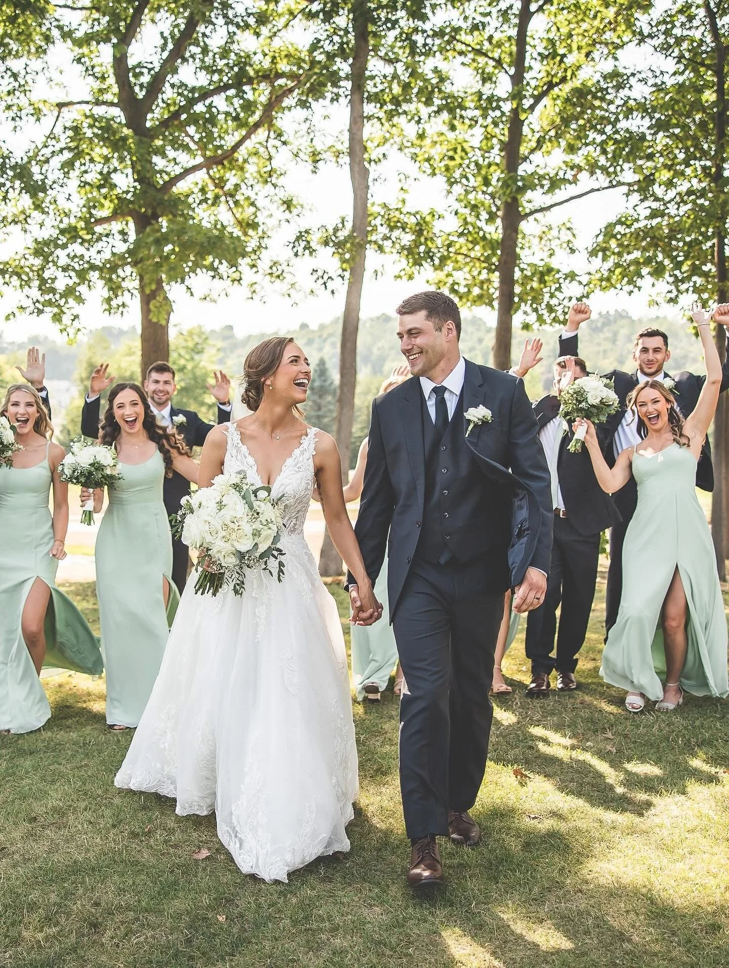 A newlywed couple walking hand in hand outdoors, surrounded by smiling friends and bridesmaids, with trees and sunshine in the background.