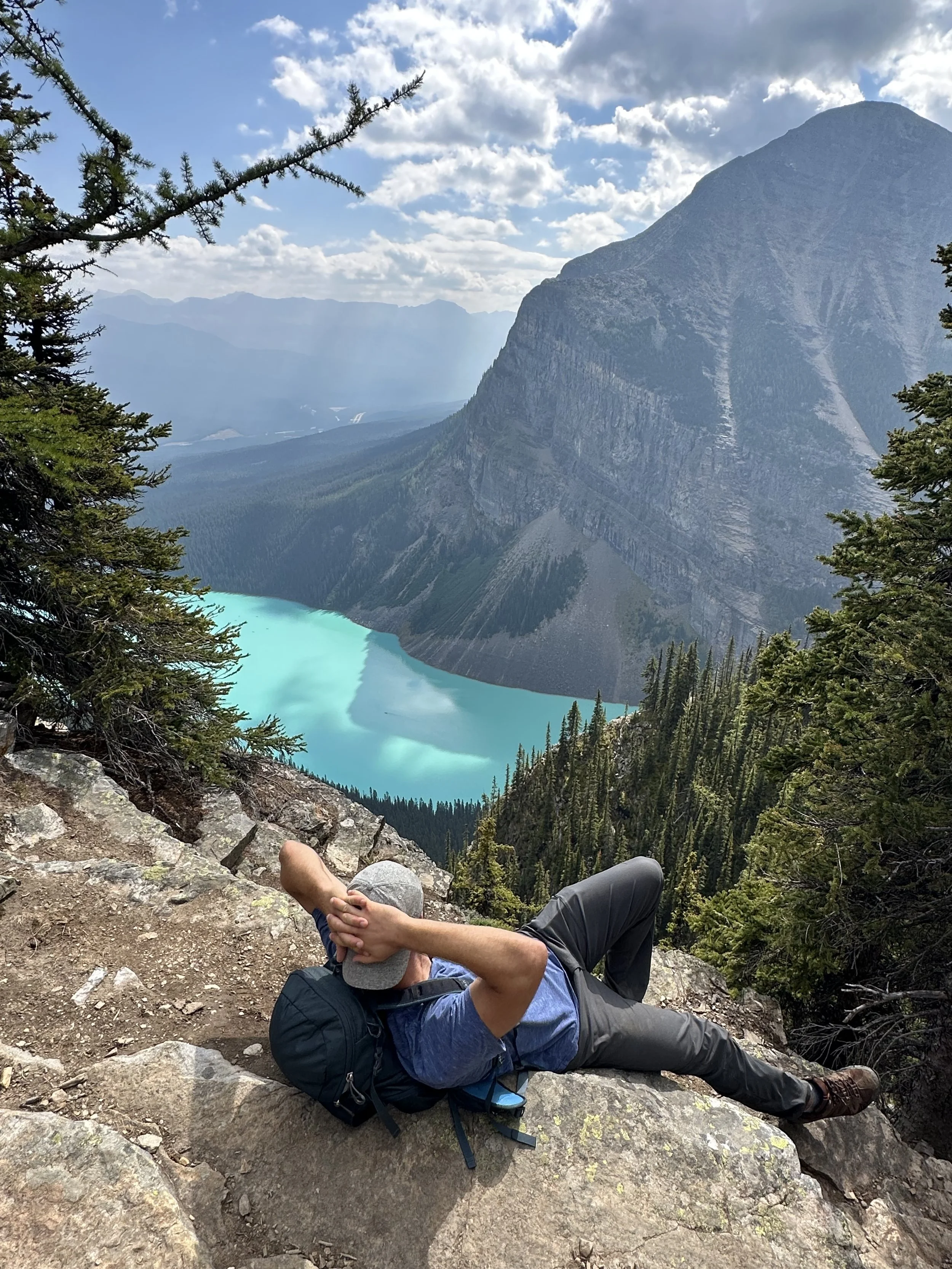 A man lying on a rocky trail resting with hands behind his head, overlooking a scenic mountain landscape with turquoise lake, green forest, tall mountains, and a partly cloudy sky.