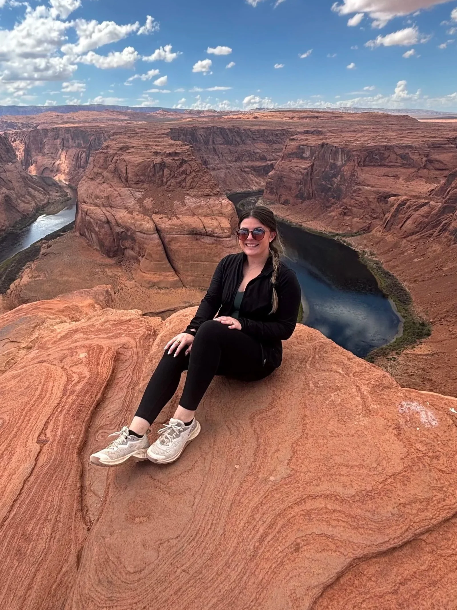 A woman sitting on a rock formation overlooking the Grand Canyon with a river winding through the canyon and a blue sky with clouds above.