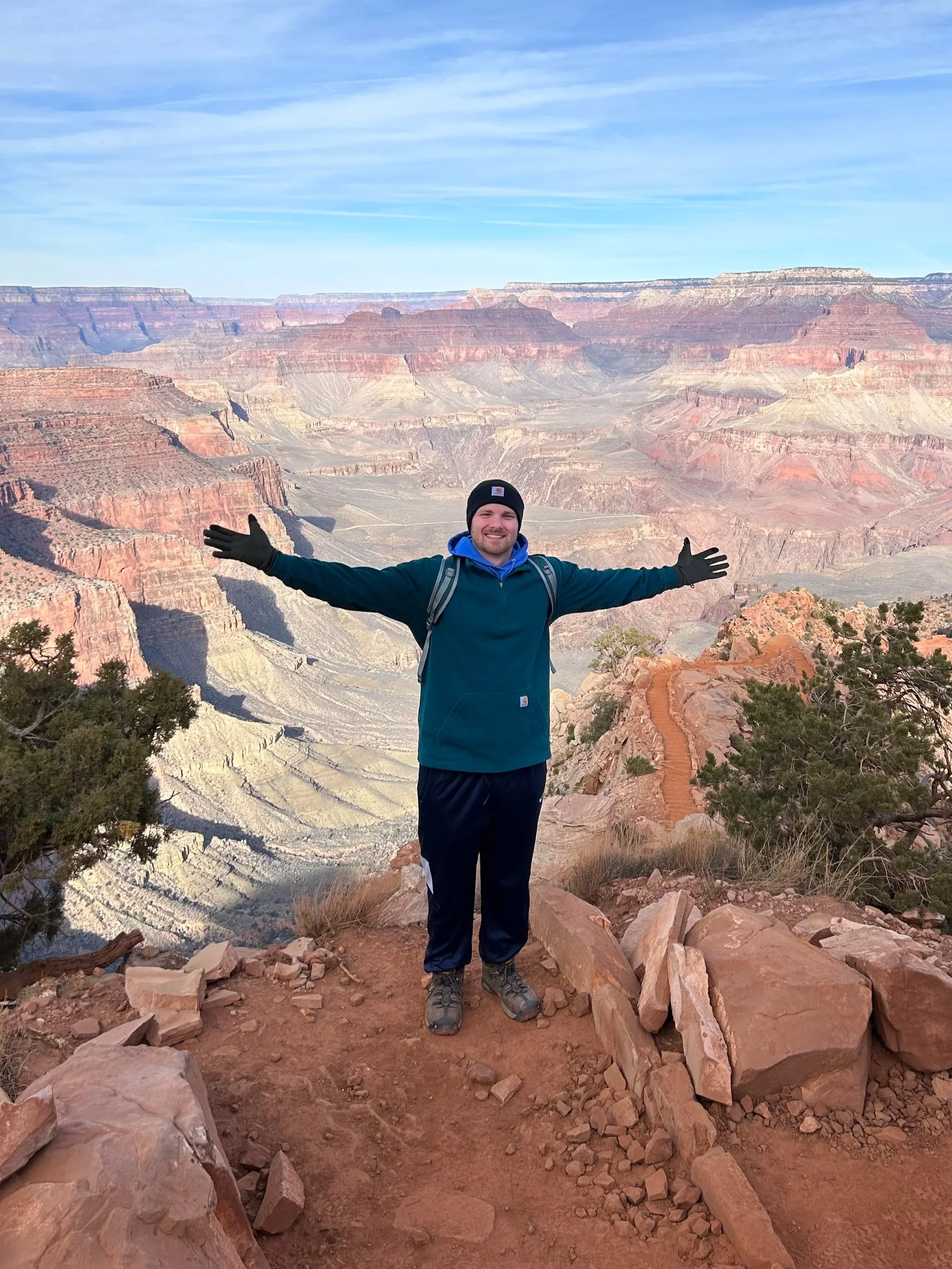 Man standing on rocky ledge with arms outstretched at Grand Canyon during daytime, with layered red and brown cliffs visible in the background.