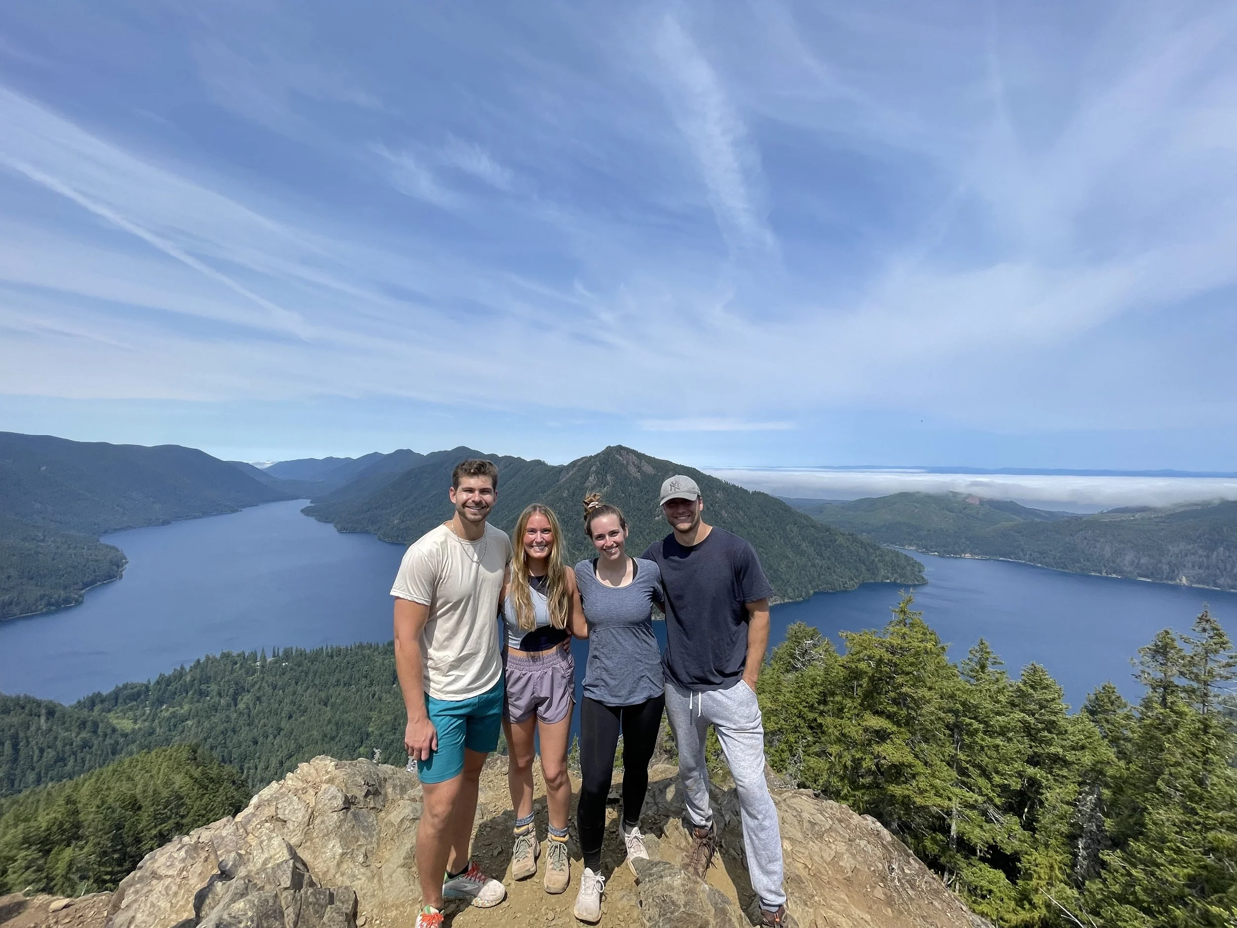 Four people standing on a rocky ledge with a scenic view of a large lake surrounded by green mountains and a blue sky with wispy clouds.