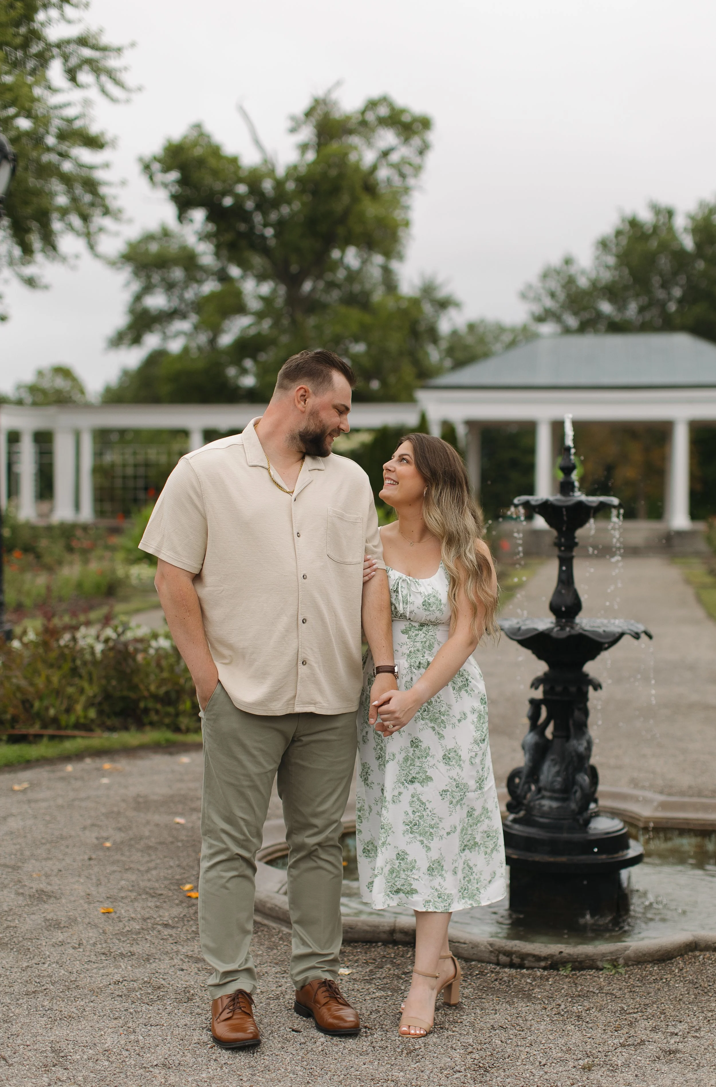 A couple standing near a fountain in a park, smiling at each other during daytime with trees and a white pavilion in the background.