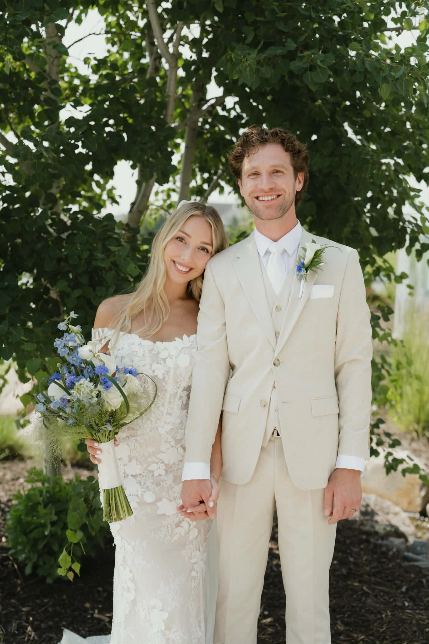 A bride and groom holding hands and smiling outdoors in front of green trees. The bride has long blonde hair and is wearing a white lace wedding dress, holding a bouquet of blue and white flowers. The groom has curly brown hair and is wearing a cream