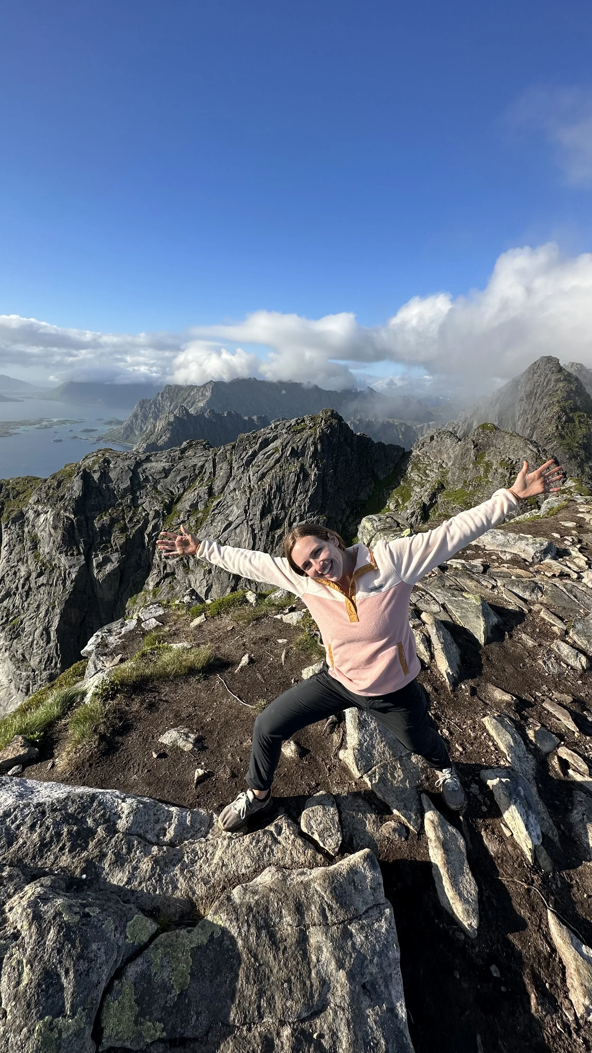 A woman celebrating on a rocky mountain trail with a panoramic view of mountains, water, and clouds in the background.