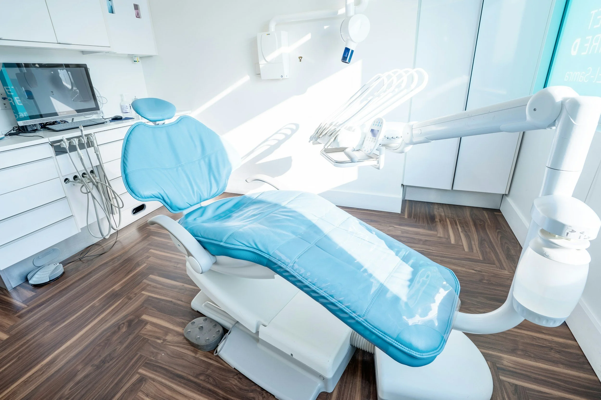 A modern dental office examination room with a blue dental chair, dental equipment, a computer, and bright natural lighting.