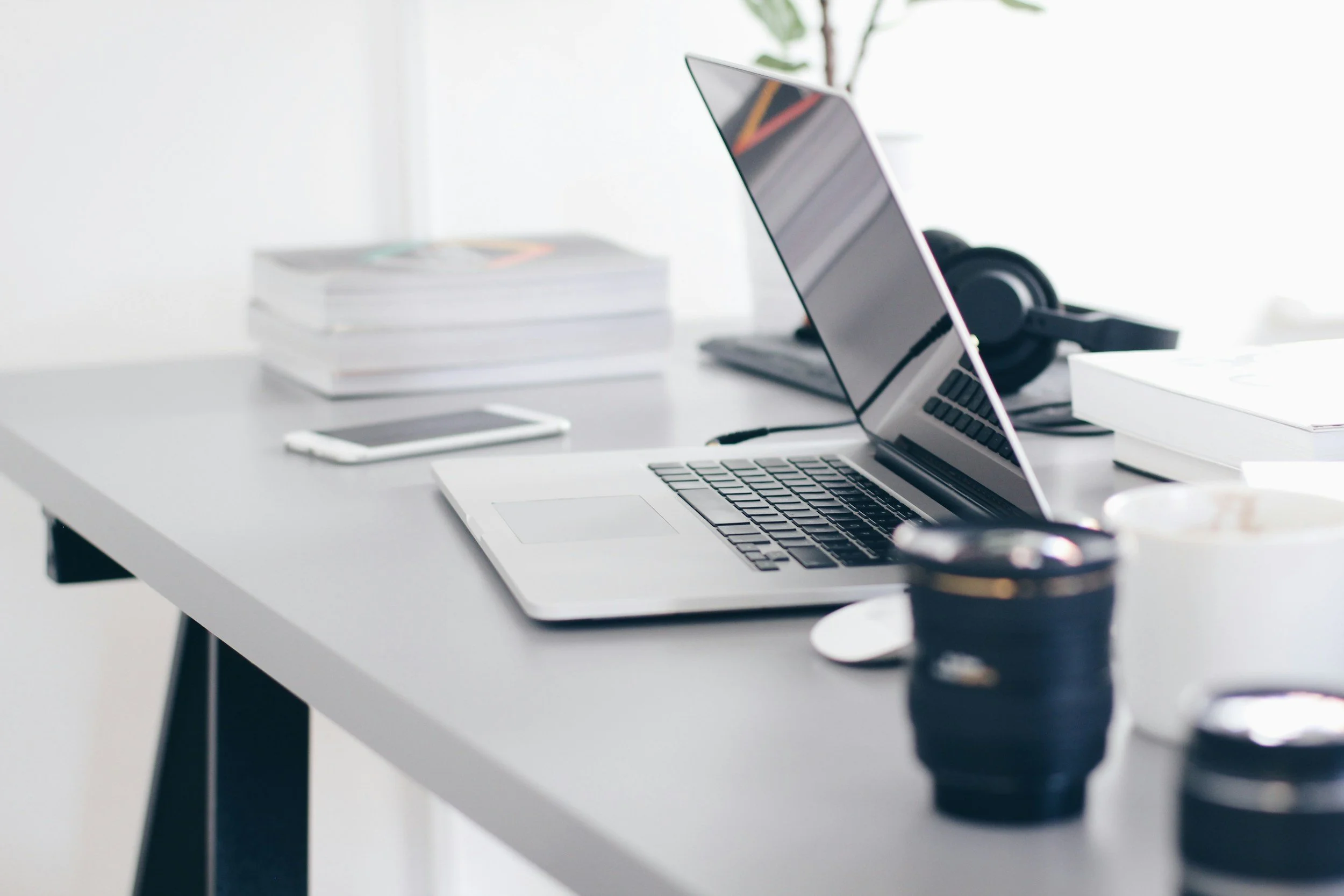 A white desk with a laptop, a smartphone, a pair of black headphones, and some stacked books in a well-lit room with a window and plants.