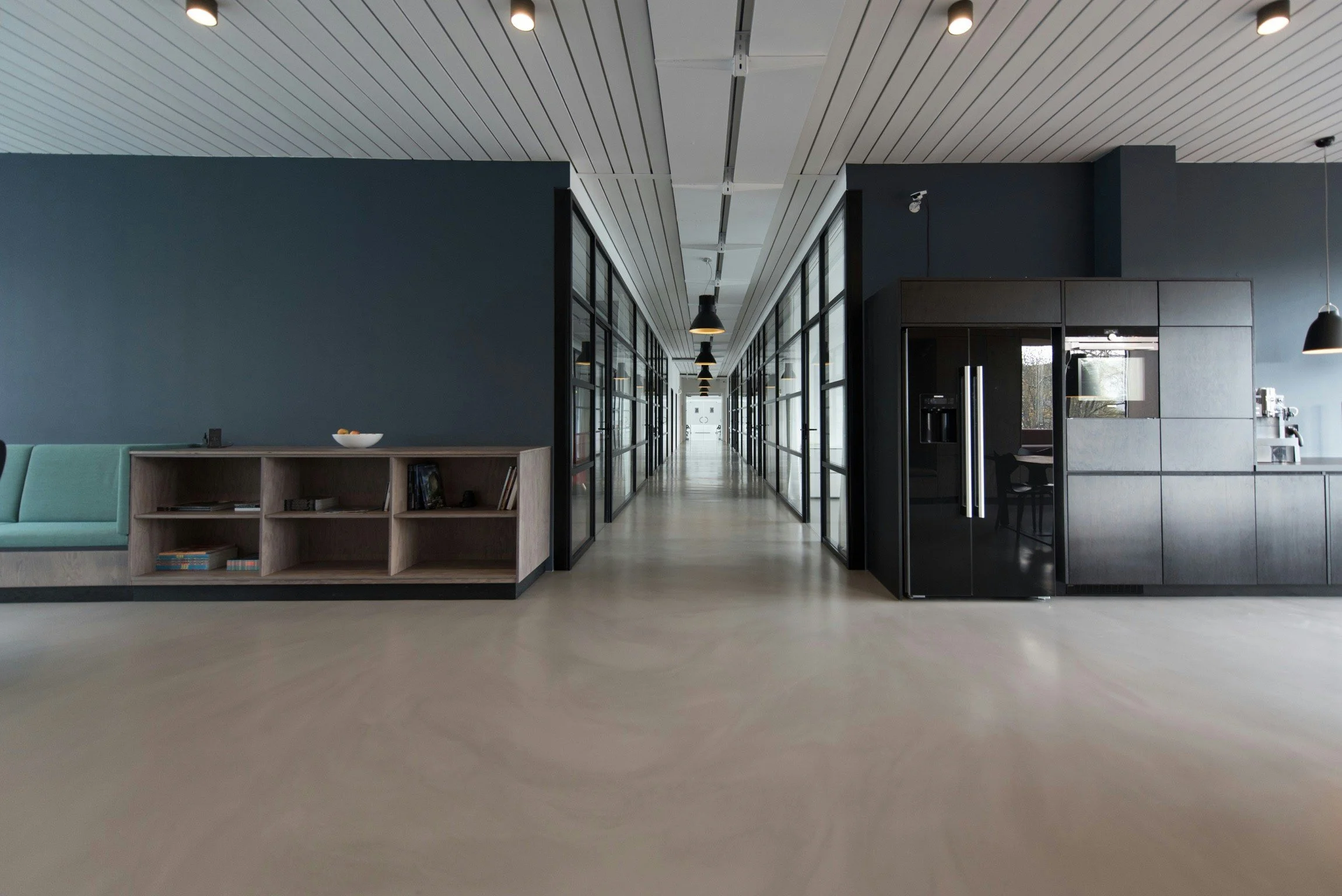 Modern interior office or communal space with a hallway featuring glass-paneled doors on both sides, black pendant lights hanging from a white ceiling with linear paneling, a partial view of a seating area with a light green sofa, and a black double-door refrigerator on the right.