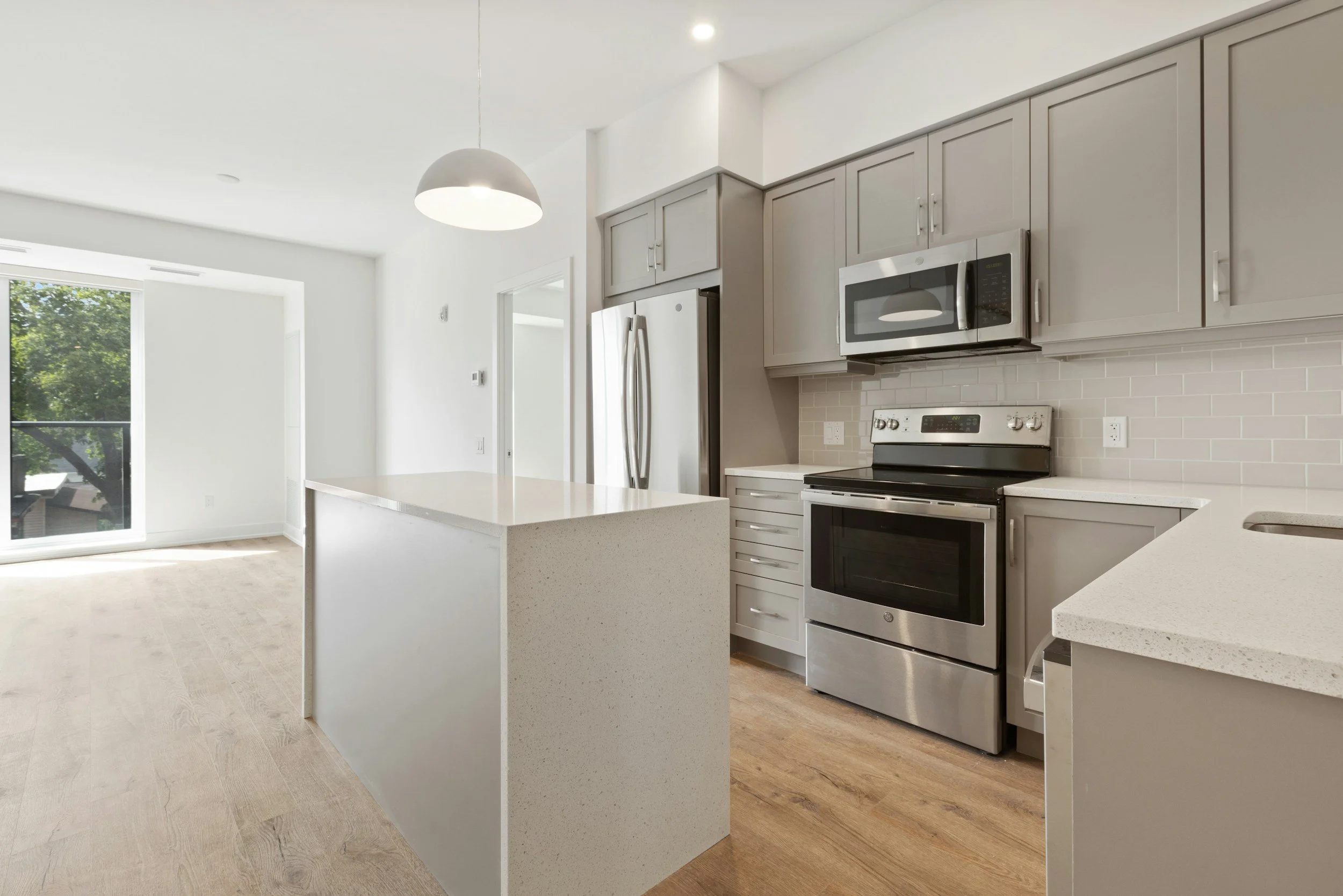 Modern kitchen with stainless steel appliances, gray cabinets, white countertops, and light wood flooring, adjacent to a bright living space with large sliding glass door.