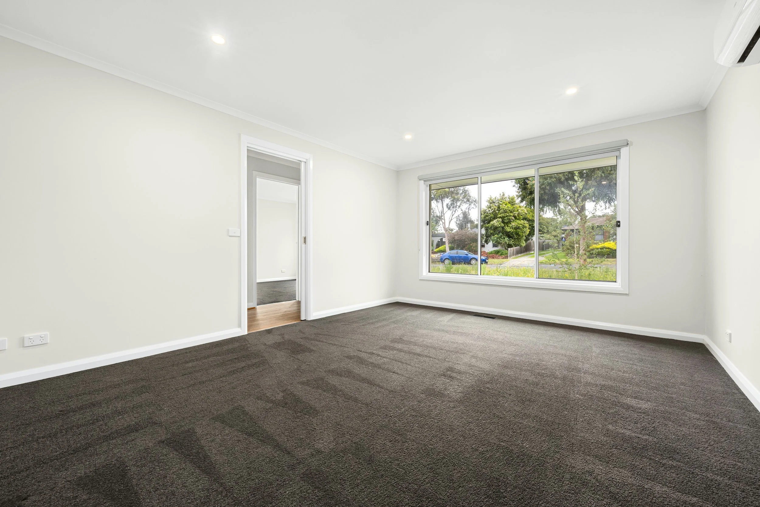 Empty room with white walls, large window with view of trees and street, dark gray carpet, air conditioning unit, and doorway to another room.