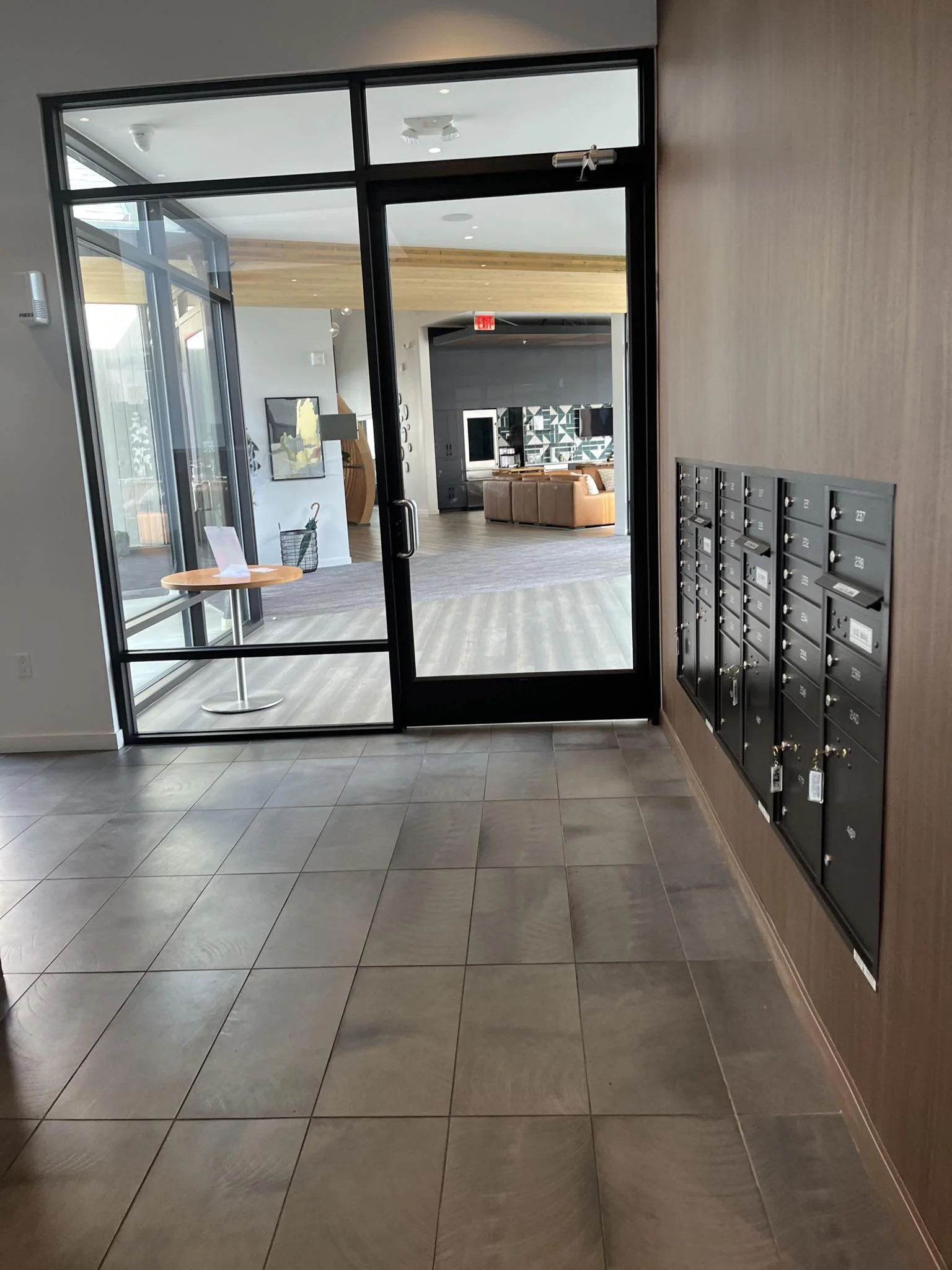 View through glass door into lobby or common area of building with mailboxes on right wall, small round table on left with paper and basket, and seating area in background with wooden ceiling and decorative wall art.