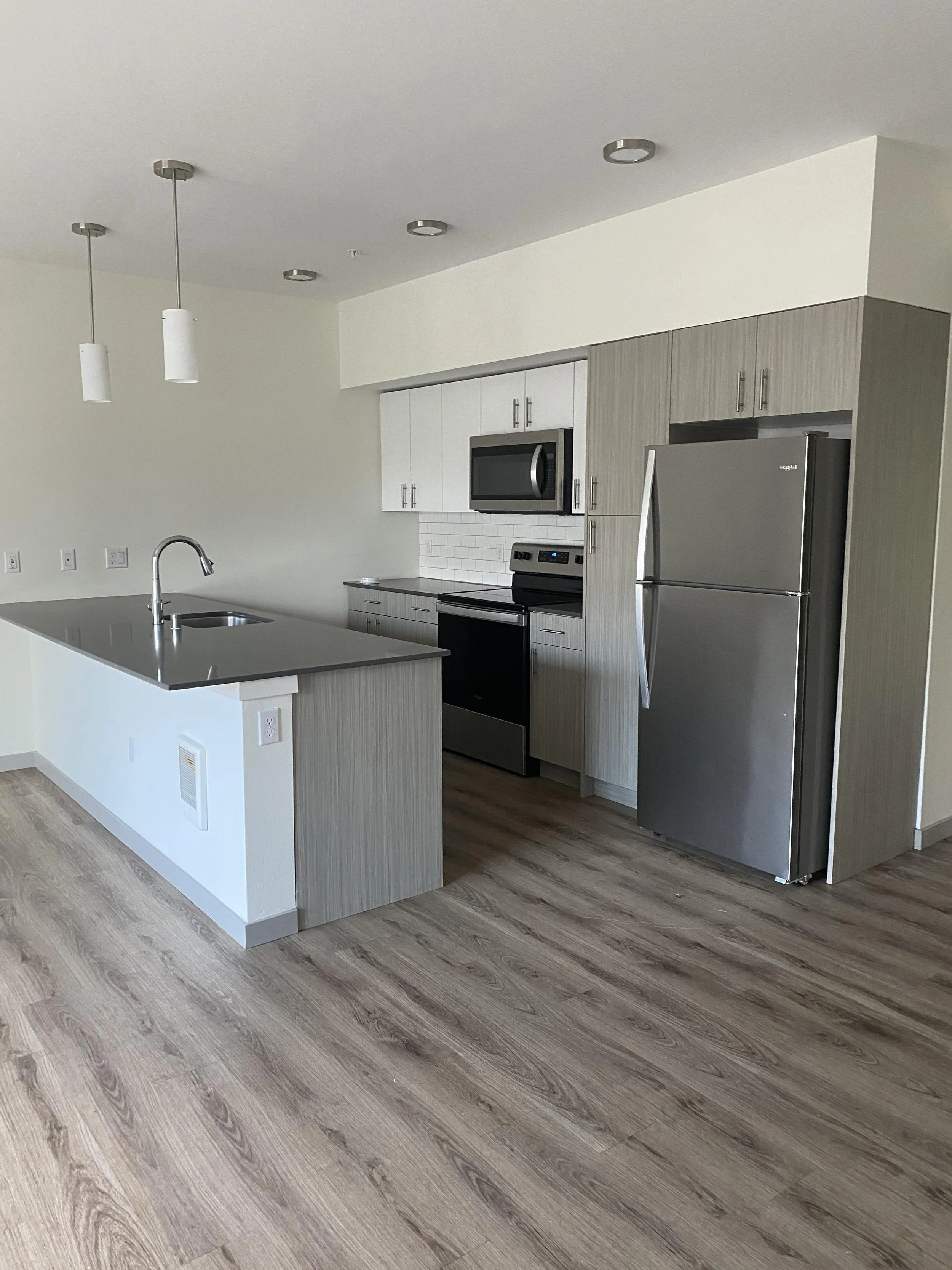 Modern kitchen with gray and white cabinetry, stainless steel refrigerator, microwave, black stove, kitchen island with sink, pendant lighting, wood flooring, and white wall with electrical outlets.
