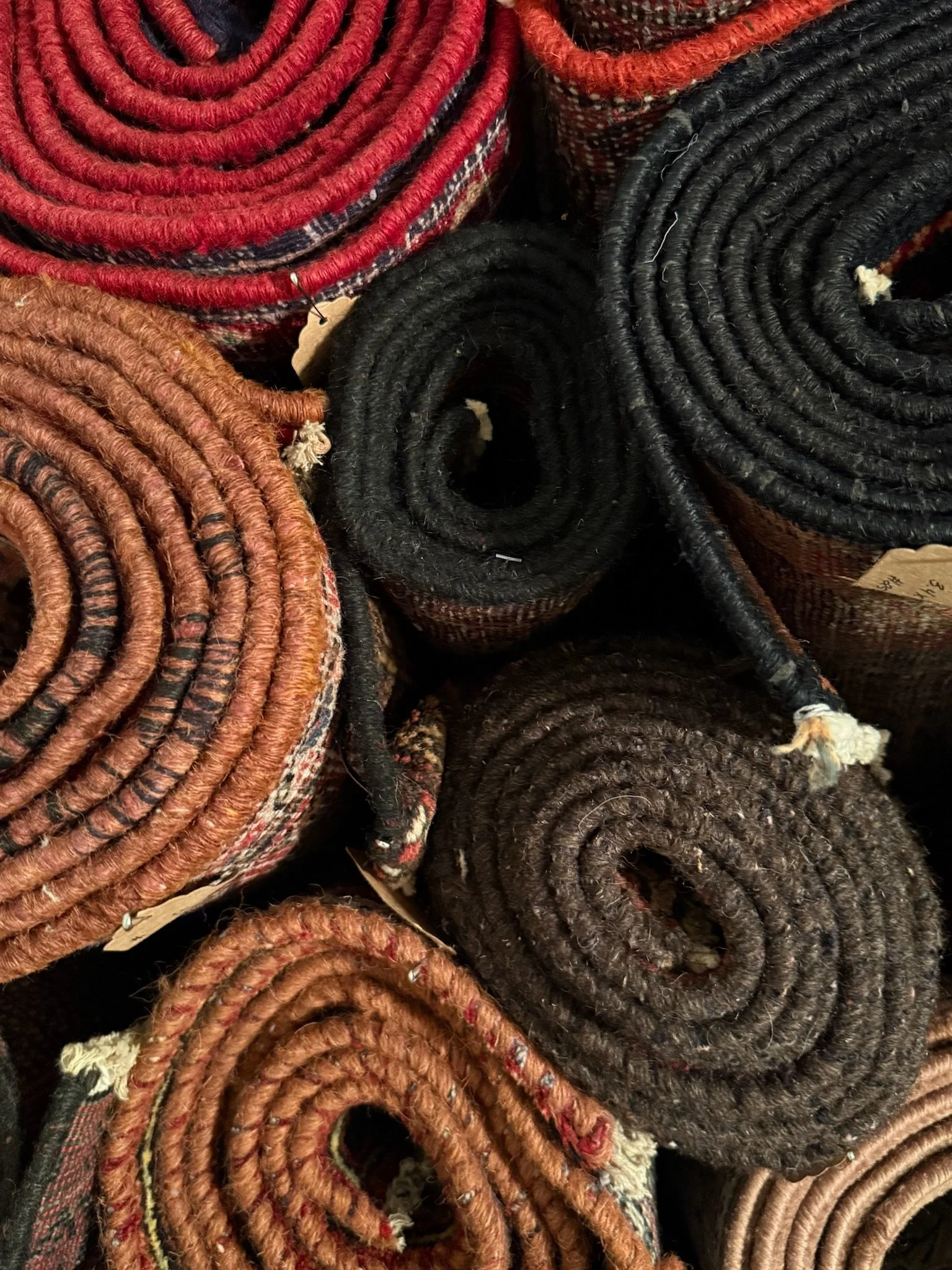 Bunches of coiled, rolled-up braided vintage handmade one-of-a-kind rugs in various colors including red, black, brown, and tan.