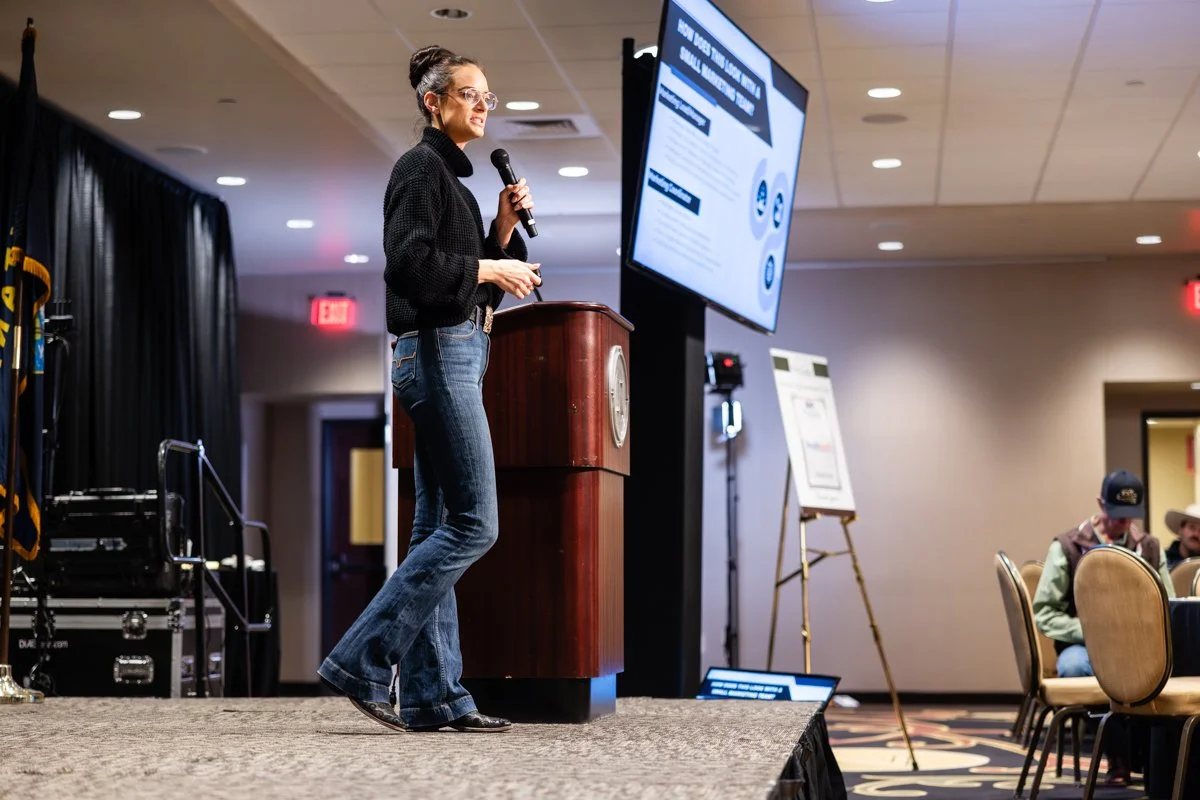 Woman giving a presentation at a conference, standing on stage with a microphone, in front of a large screen displaying slides, in a conference room with audience members seated at tables.
