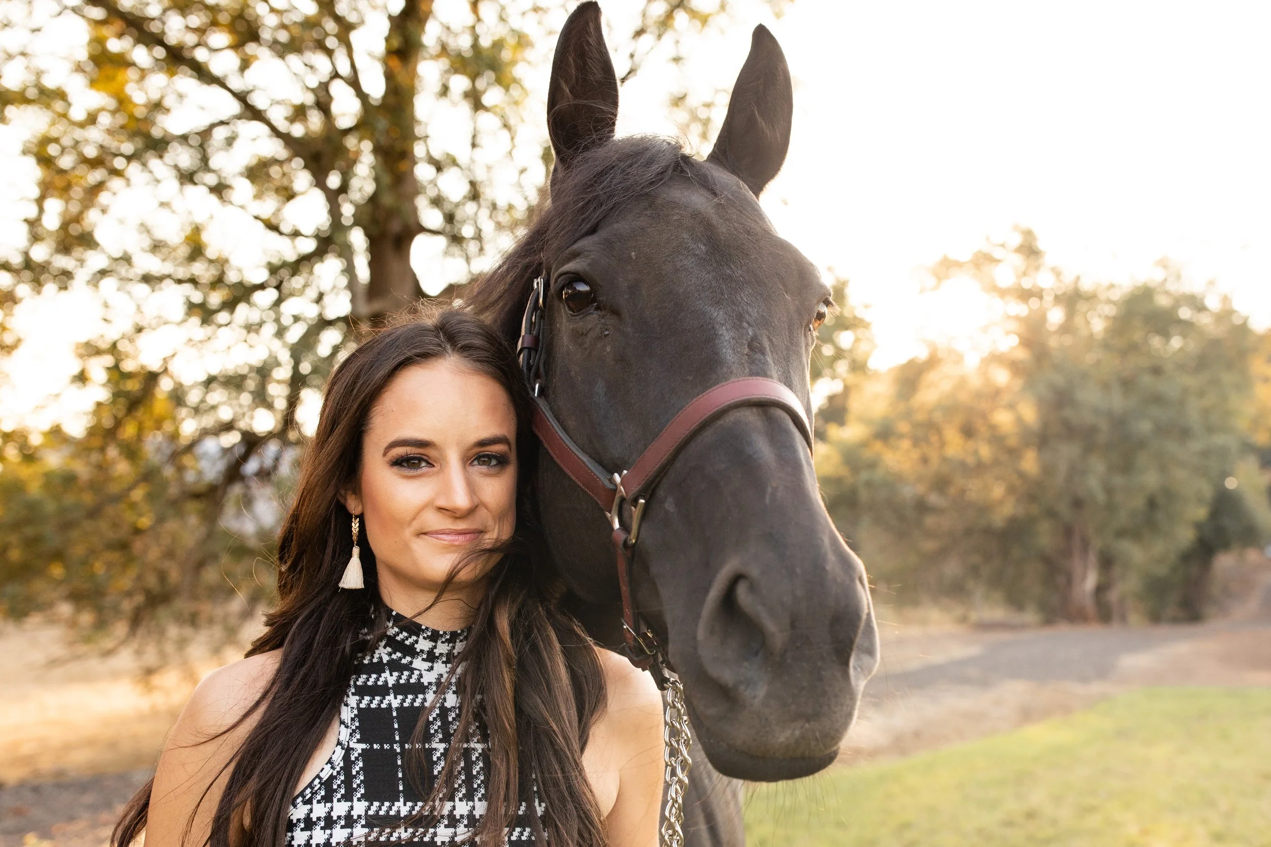A woman with long dark hair and earrings standing next to a black horse, with trees and a sunset in the background.