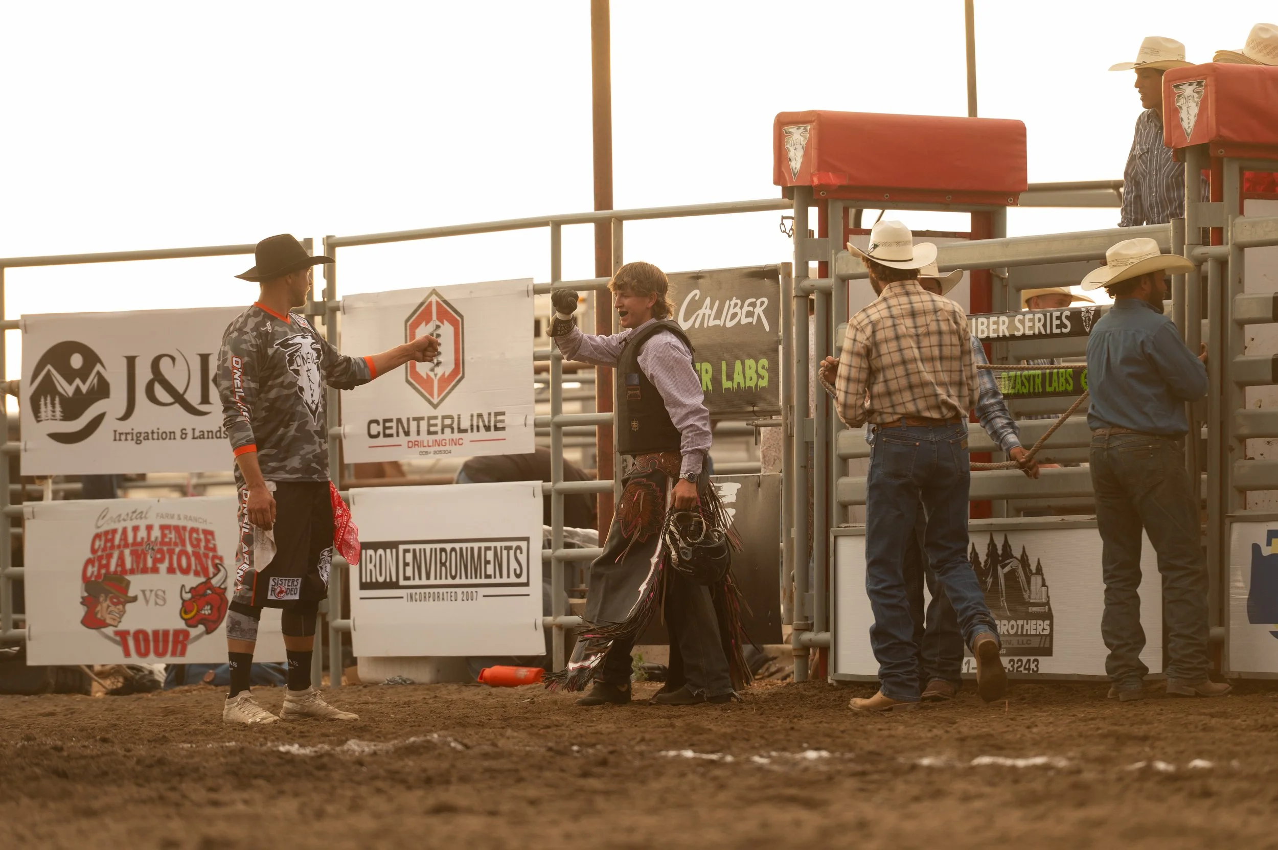 People at a rodeo event standing in the arena near the gate with sponsor banners in the background, dirt arena visible in the foreground.