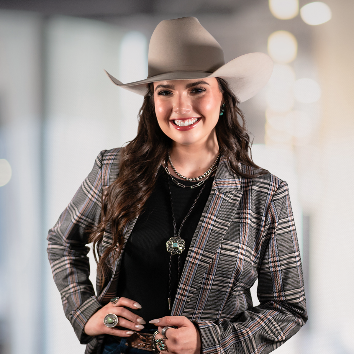 A smiling woman wearing a grey cowboy hat, a plaid blazer, and various jewelry, standing indoors with a blurred background of lights.