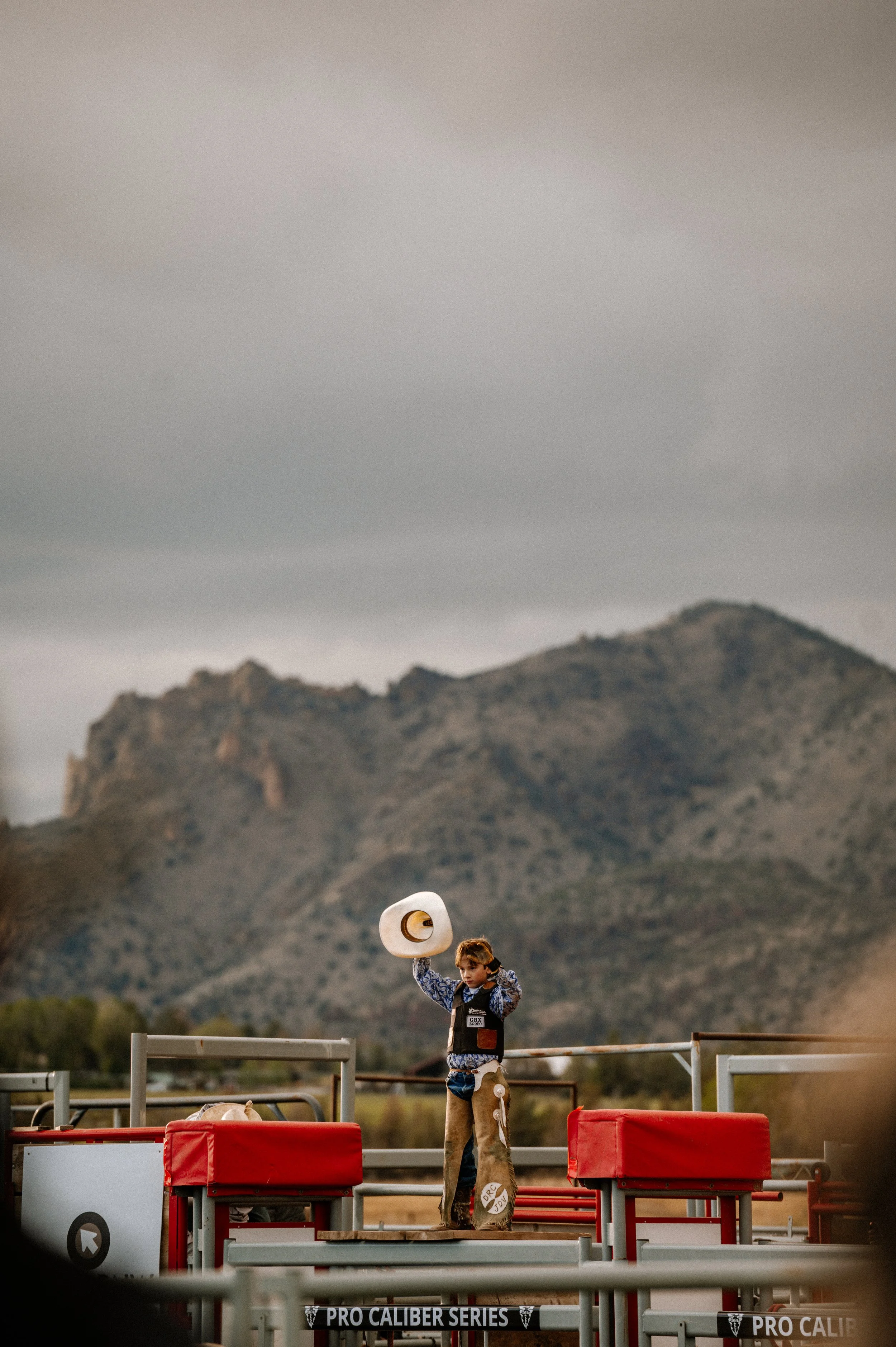 A young boy holding a cowboy hat above his head standing on a rodeo arena.
The background shows mountains and cloudy sky, with rodeo equipment in the foreground.