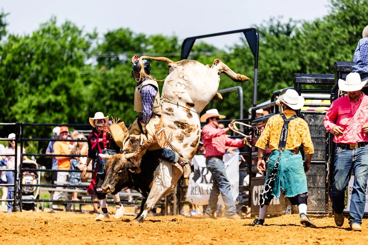 A rodeo event where a cowboy is riding a bucking bull, surrounded by rodeo staff and spectators, with some people wearing cowboy hats and western attire.