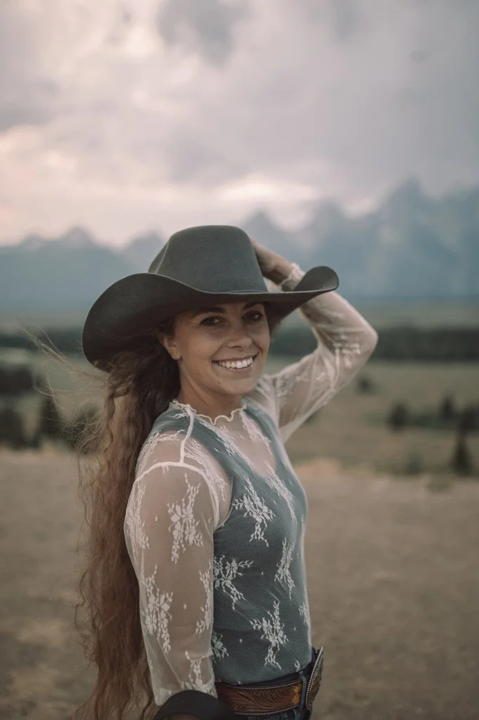 A woman wearing a gray cowboy hat, a sheer long-sleeved lace top with floral patterns, smiling with her hand on the hat, standing outdoors in a vast landscape with cloudy sky and mountains in the background.