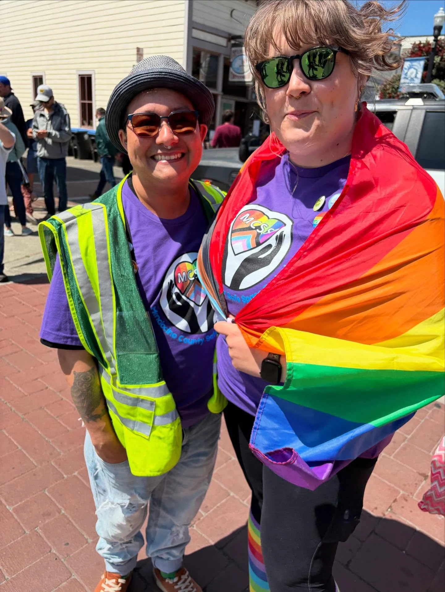 Two volunteers stand together at the edge of a Pride Parade smiling and showing off their MCSSP logo tee-shirts. One is wearing a security vest and the other has a rainbow pride flag draped over their shoulders.