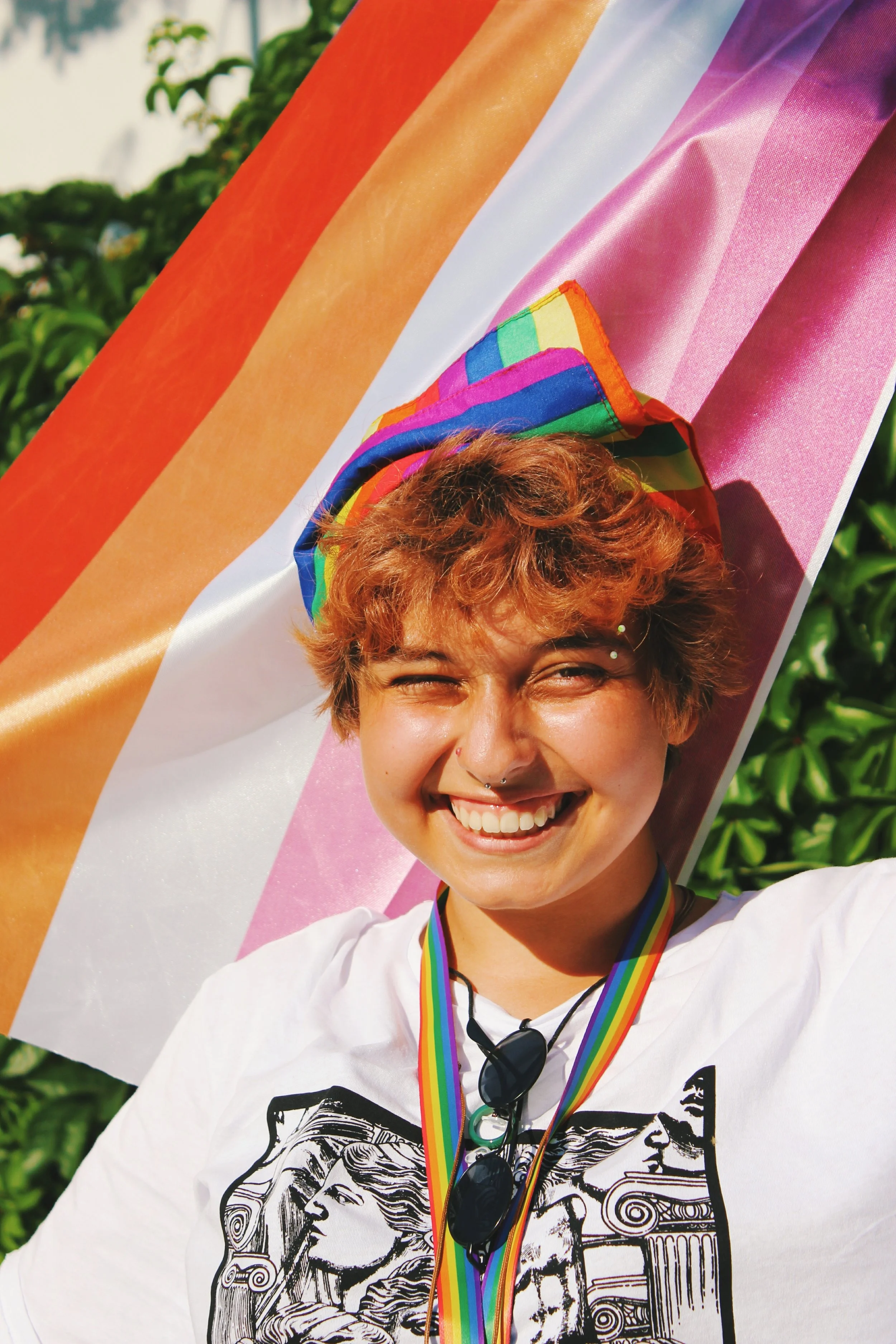 A young adult smiles while holding a multi-colored trans-flag outdoors.