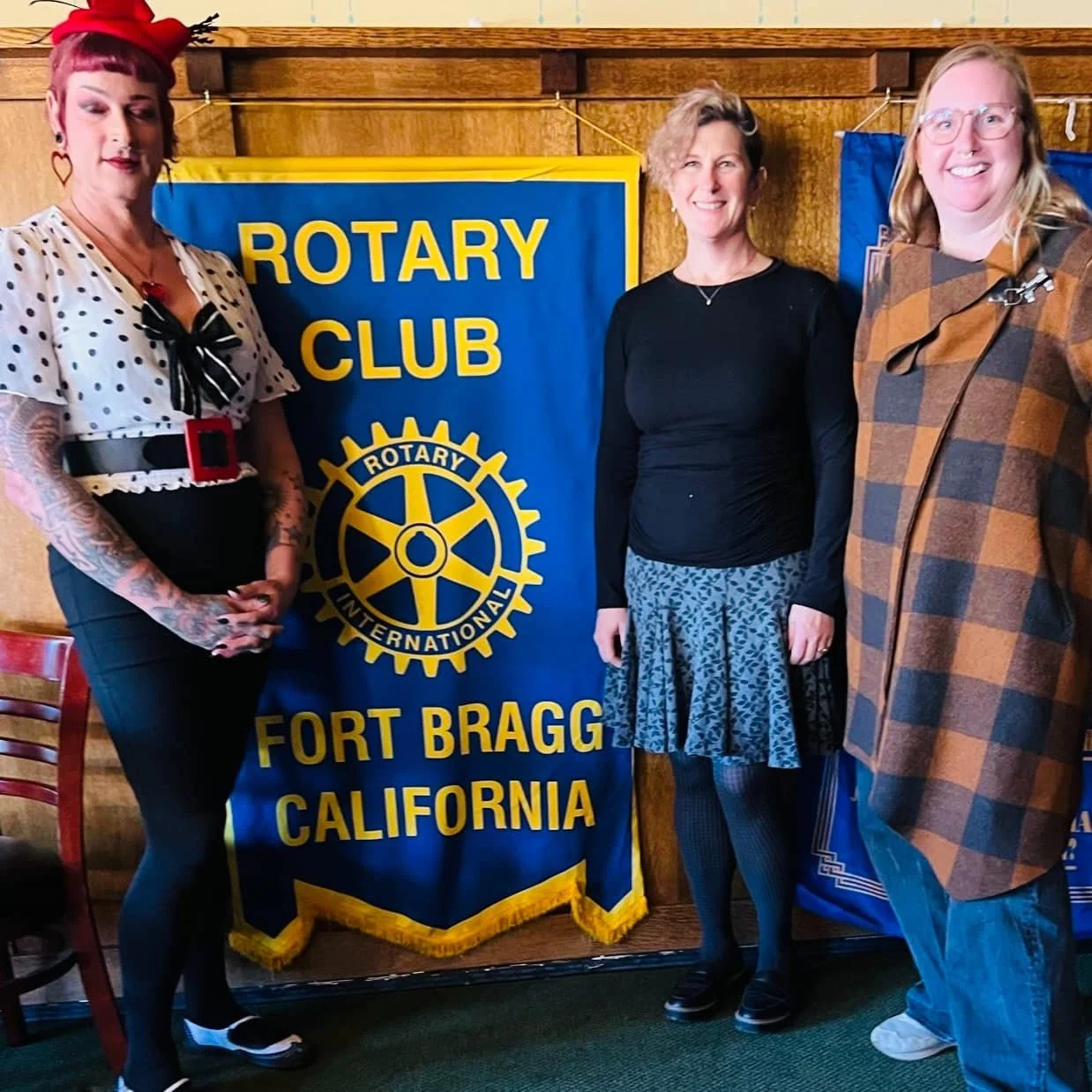Three people stand together posing confidently in front of a blue and gold banner for the Rotary Club of Fort Bragg California