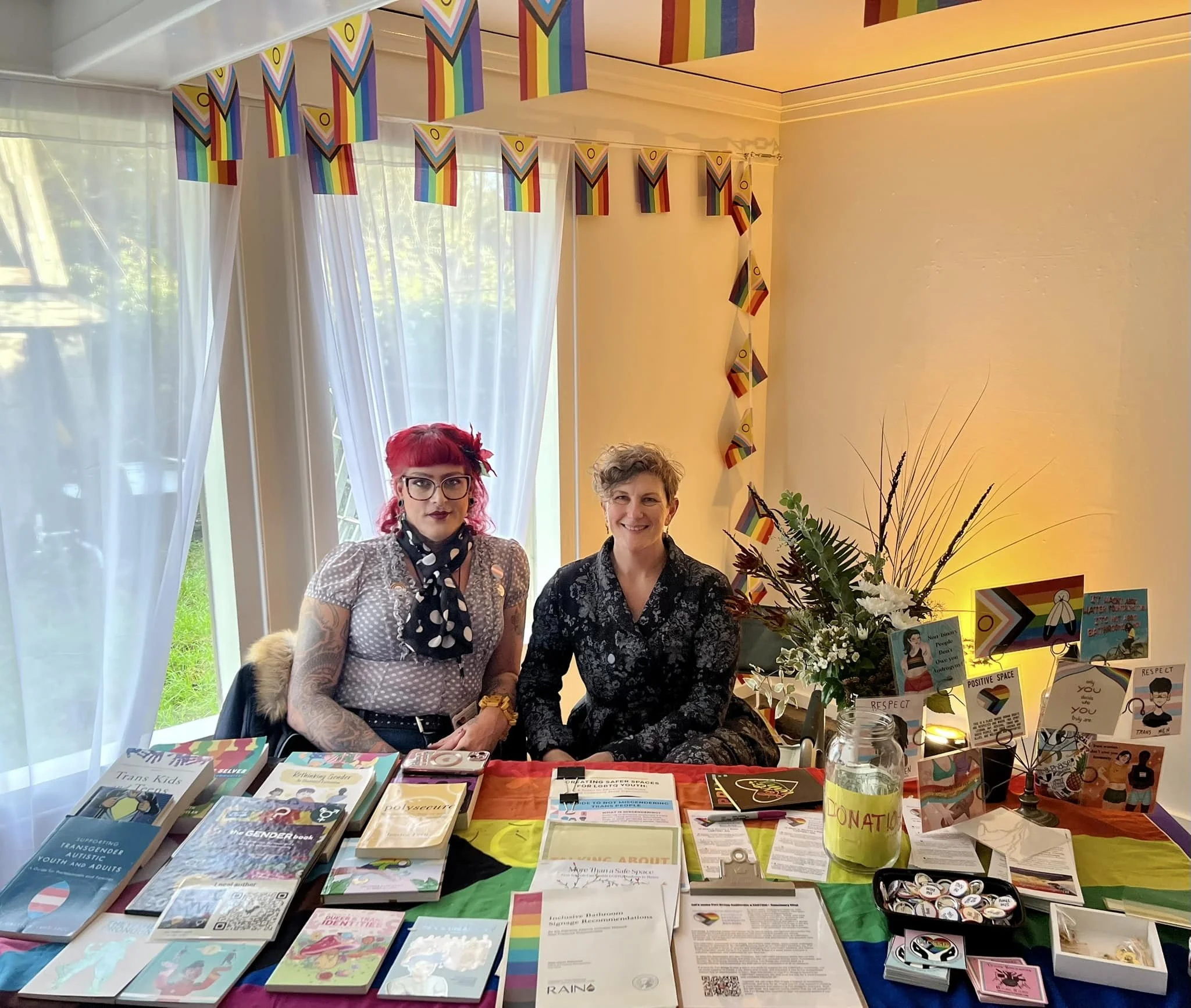 A transgender woman and their non-binary partner are seated at a table displaying educational materials on Cultural Humility and Inclusive Practices.
