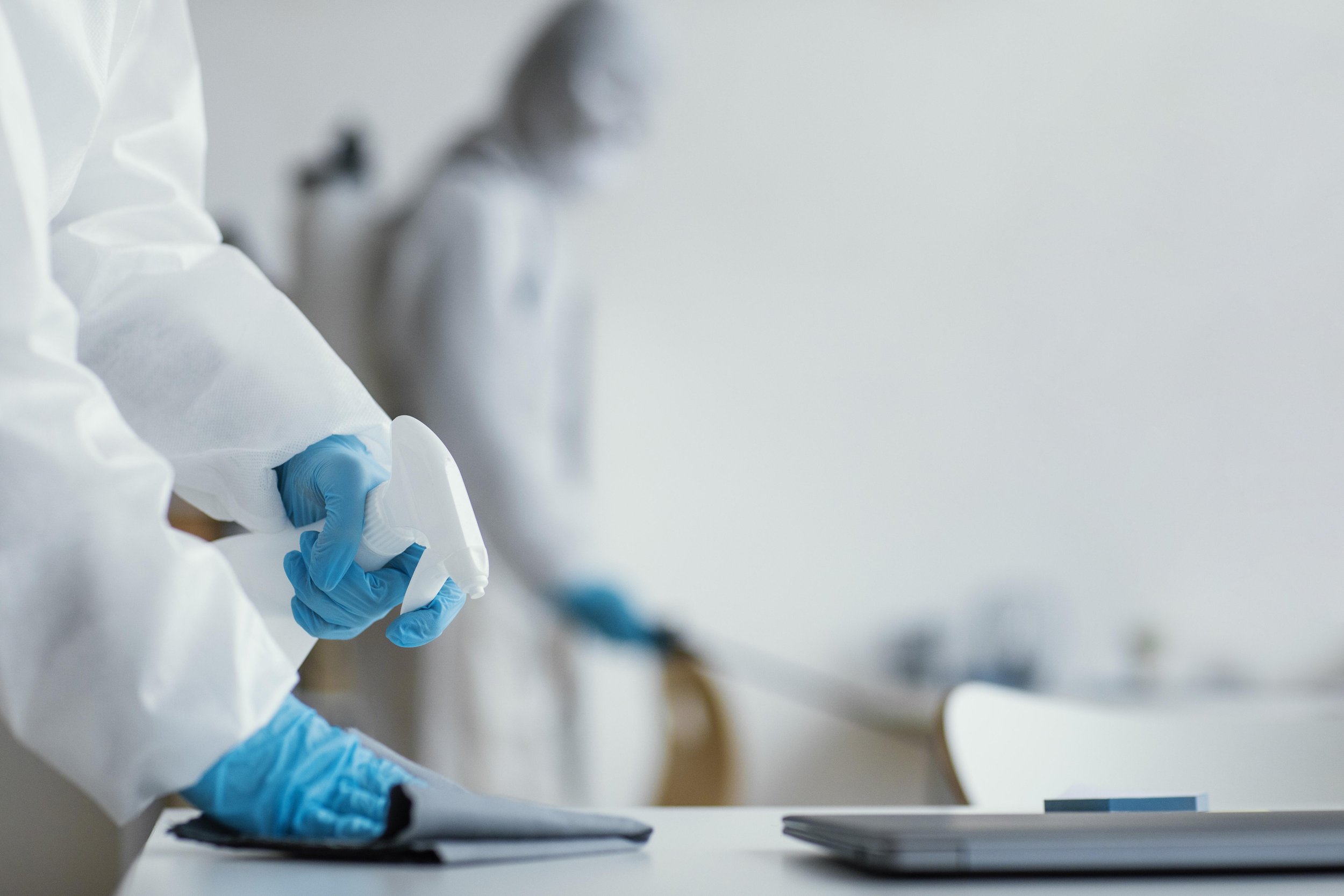 Person in a white protective suit and blue gloves holding a spray bottle, with another person in similar attire blurred in the background.