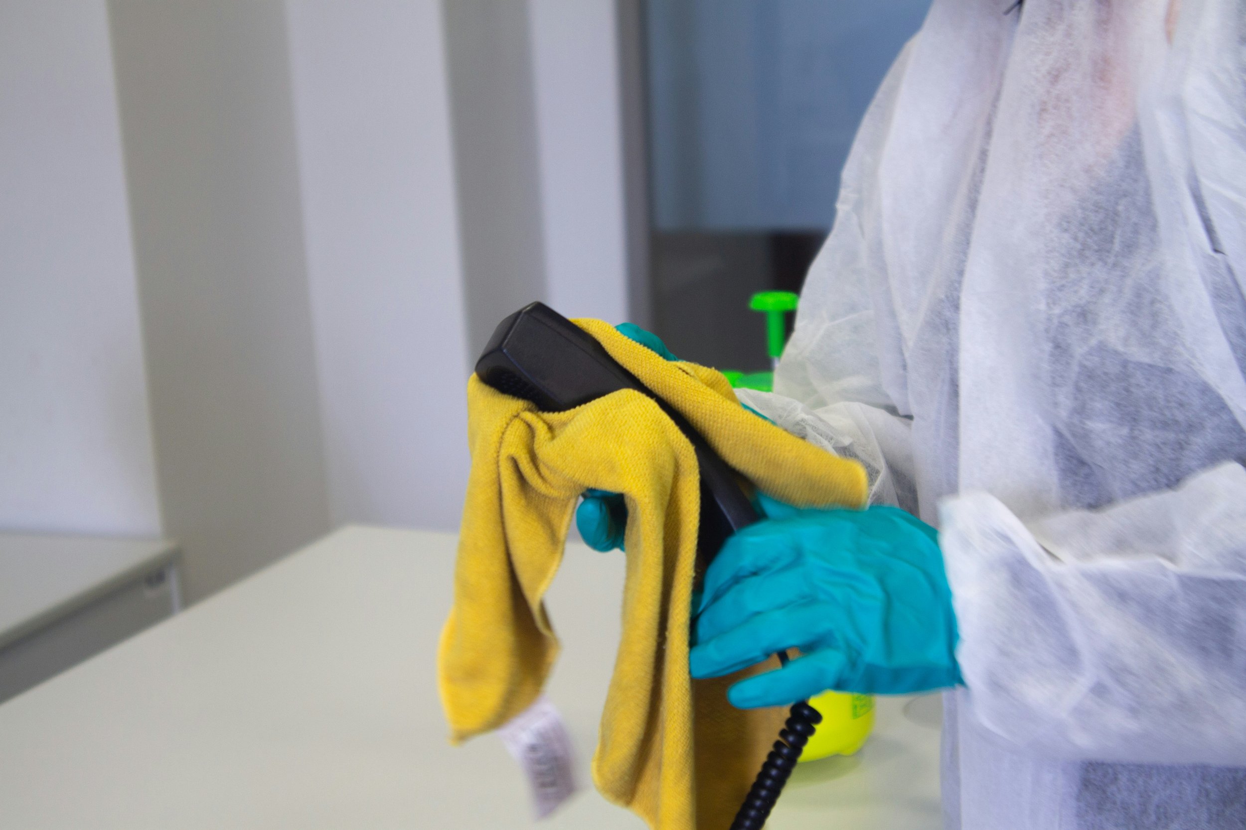 A person in protective gear, including gloves and a gown, holding a disinfectant spray and a yellow cloth in a clinical setting.