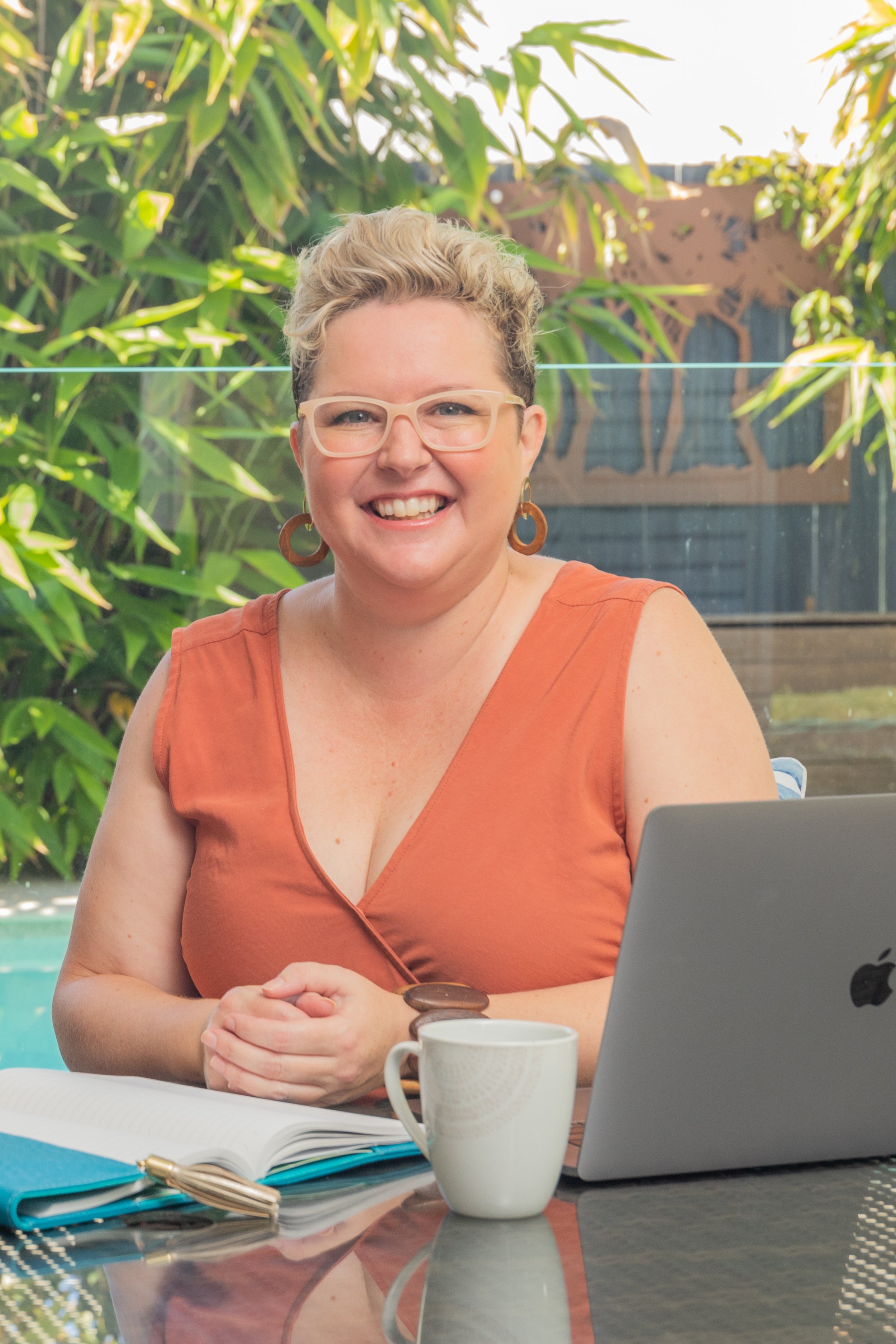 A smiling woman with short, curly blonde hair wearing glasses and large earrings, sitting at a table outdoors. There are books, a laptop, a coffee mug, and some coins in front of her. The background features green plants and a wooden fence.