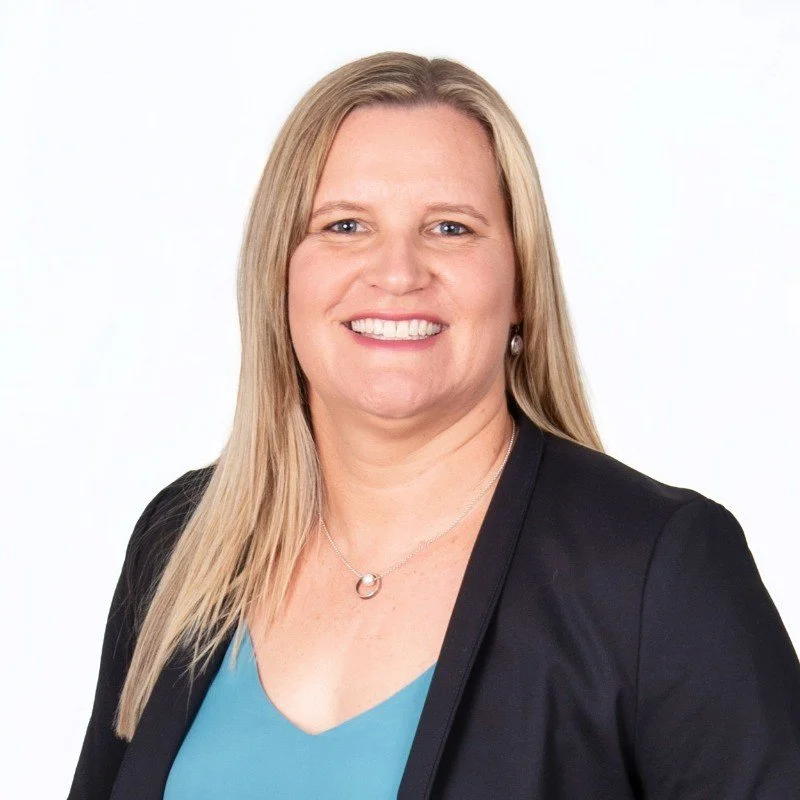 A professional woman with long blonde hair smiling, wearing a black blazer, a light blue top, and jewelry, against a plain white background.