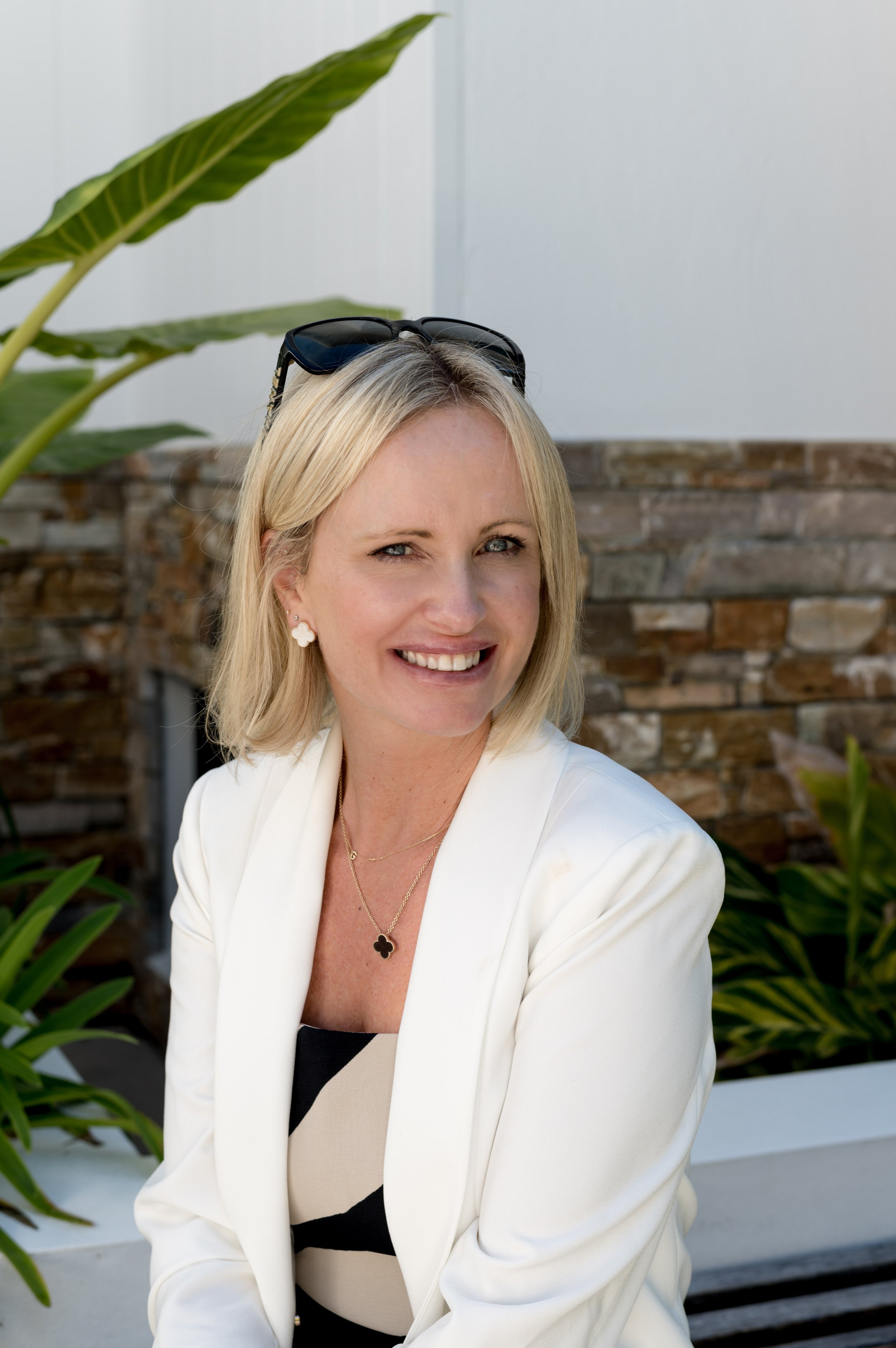 A woman with blonde hair, wearing sunglasses on her head, a white blazer, a beige and black top, and jewelry including earrings and a necklace, smiling outdoors with a background of a brick wall, a white wall, and green plants.