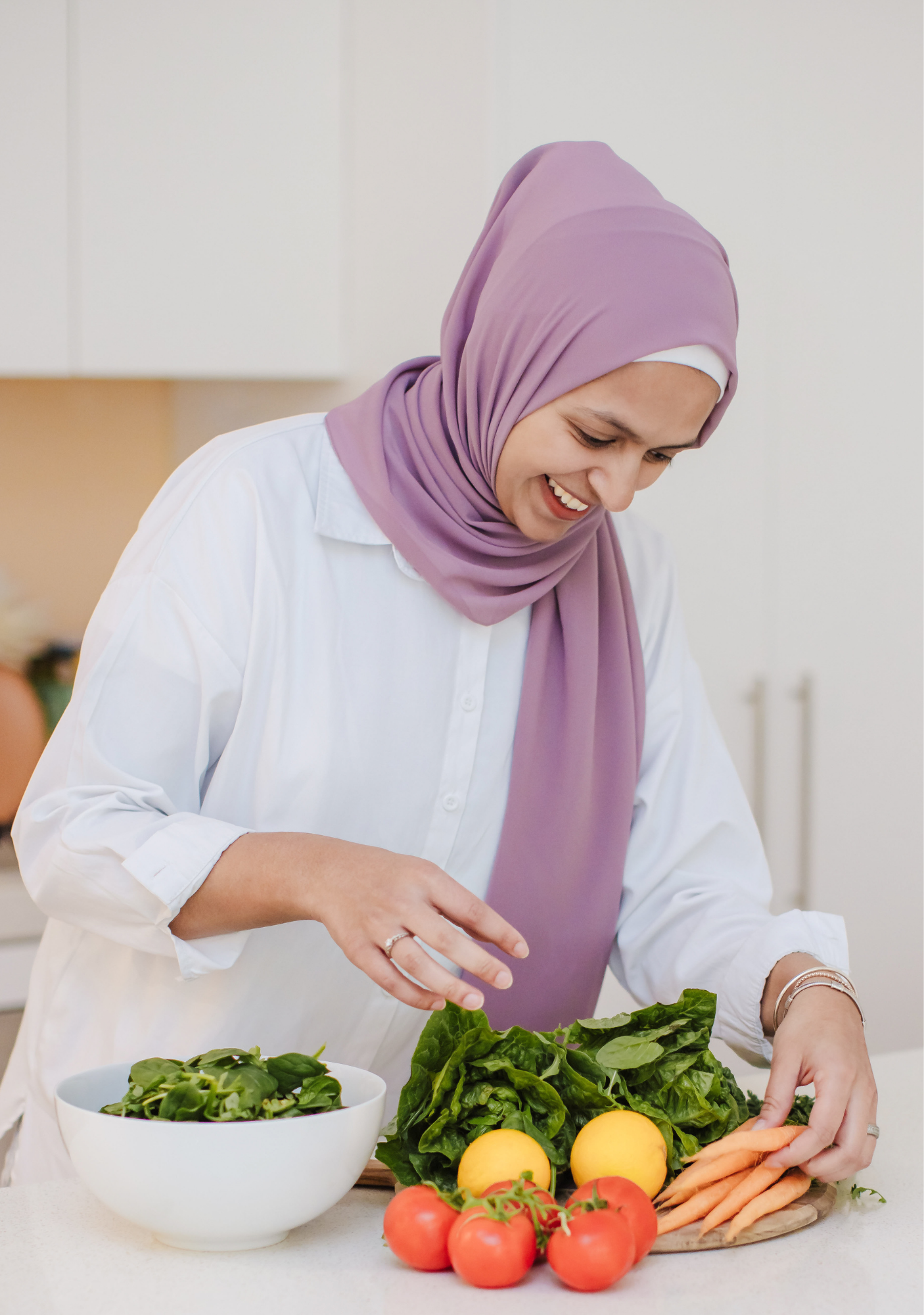 A woman wearing a lavender hijab and white shirt preparing fresh vegetables in a kitchen.