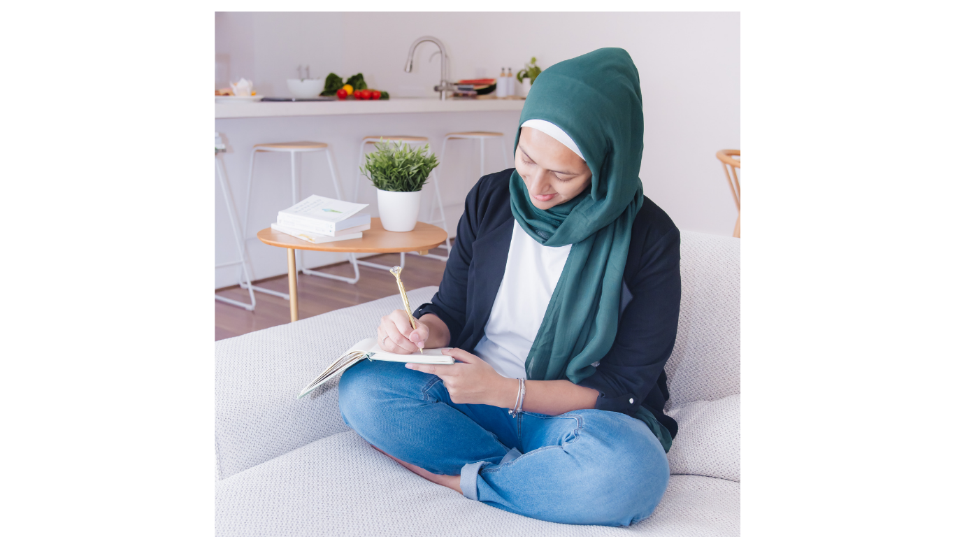 A woman wearing a green hijab and casual attire sitting on a sofa, writing in a notebook.