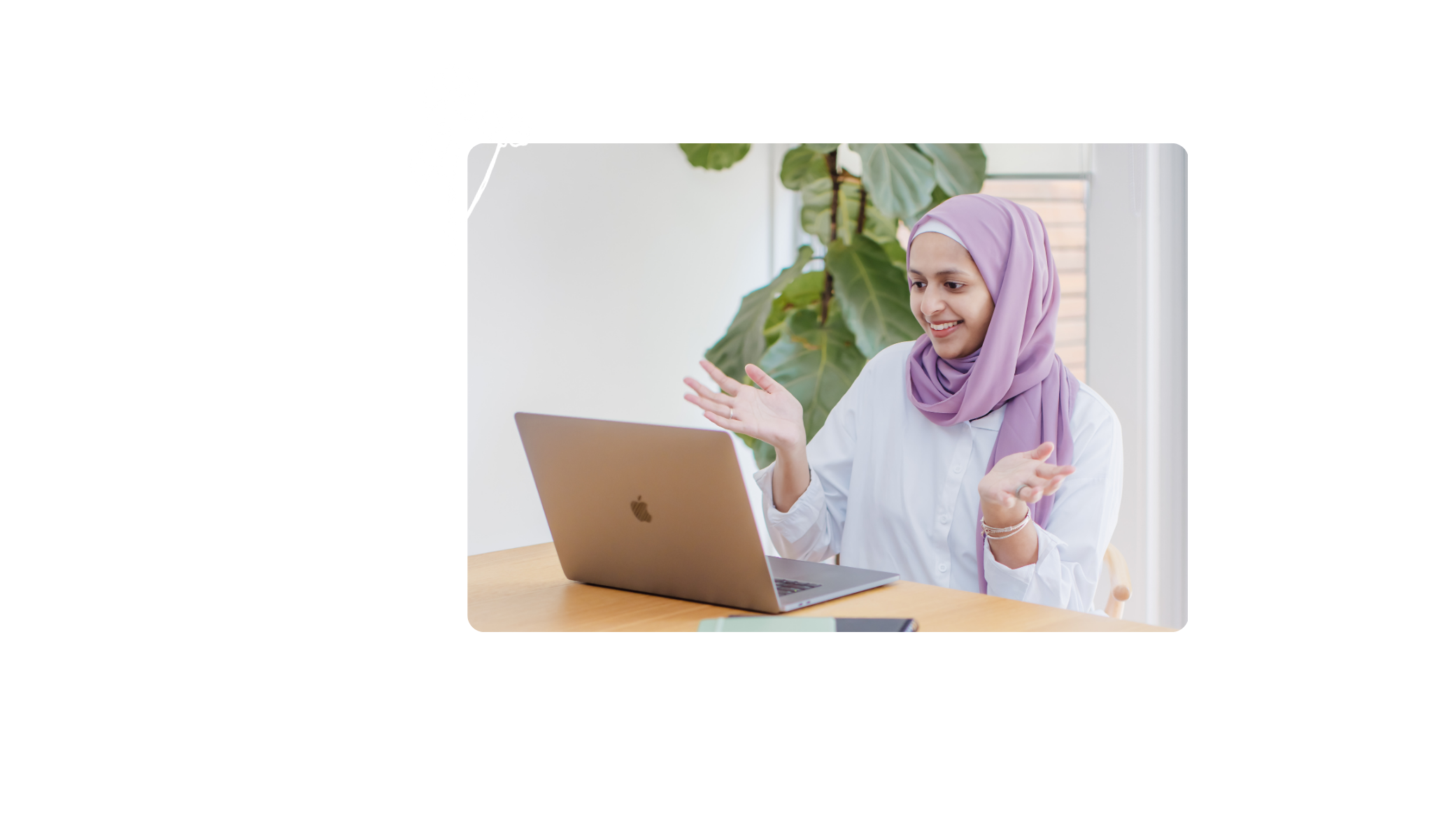 Young woman wearing a lavender hijab and white shirt smiling and gesturing with her hands while talking during a video call on her laptop at a wooden desk with a potted plant in the background.