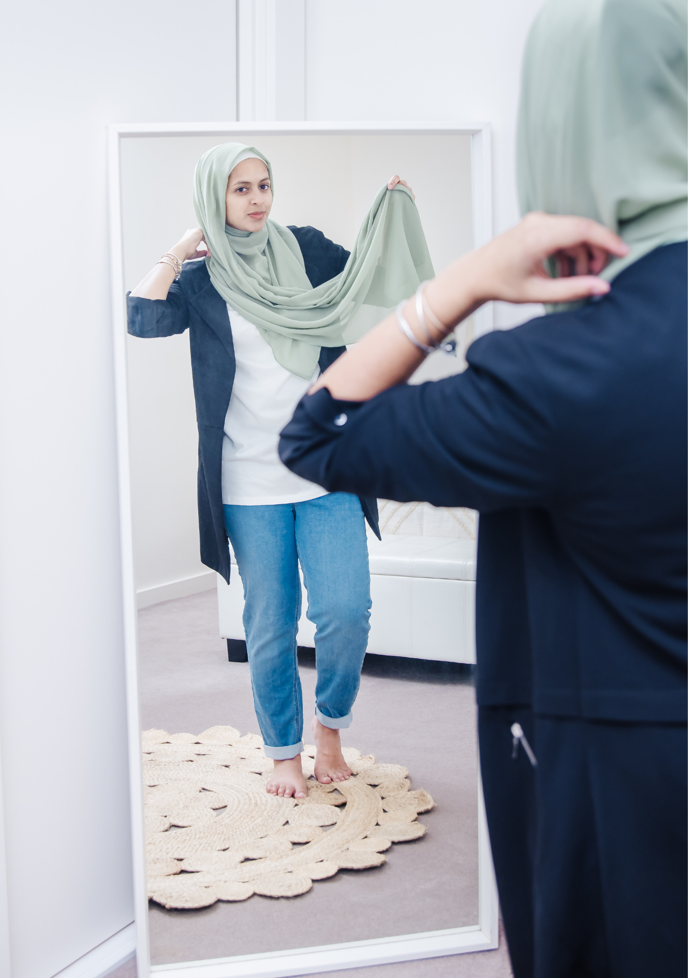 A woman trying on a green headscarf while looking in the mirror, with her hands adjusting the scarf.