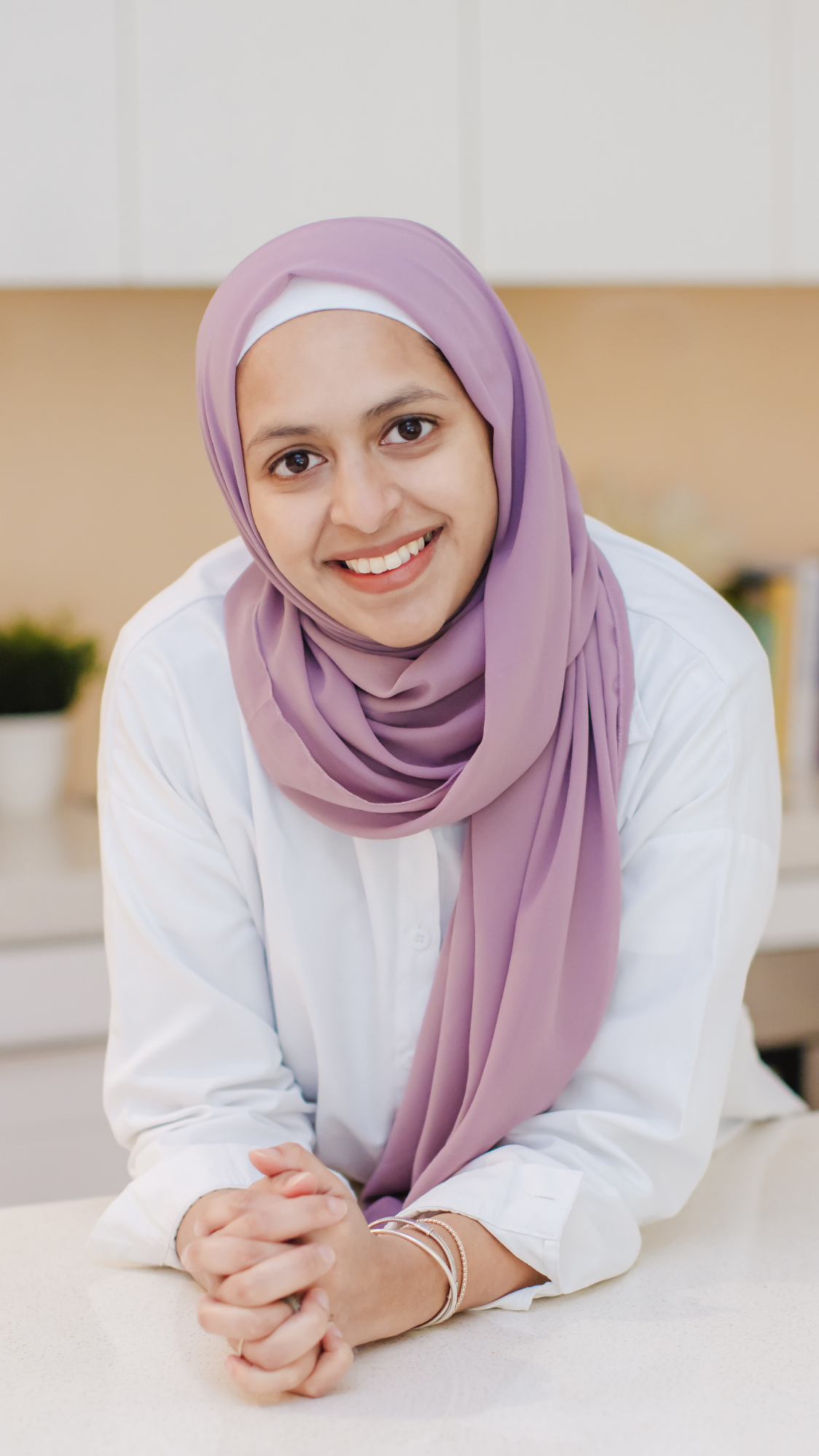 Smiling woman wearing a lilac hijab and white shirt, seated at a kitchen counter.