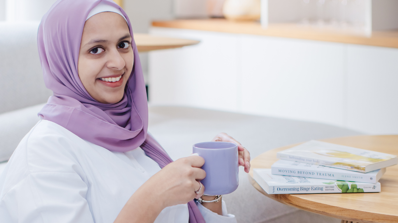 A woman wearing a purple hijab and white shirt sitting at a table holding a purple mug, with books about healing, trauma, and binge eating in front of her.