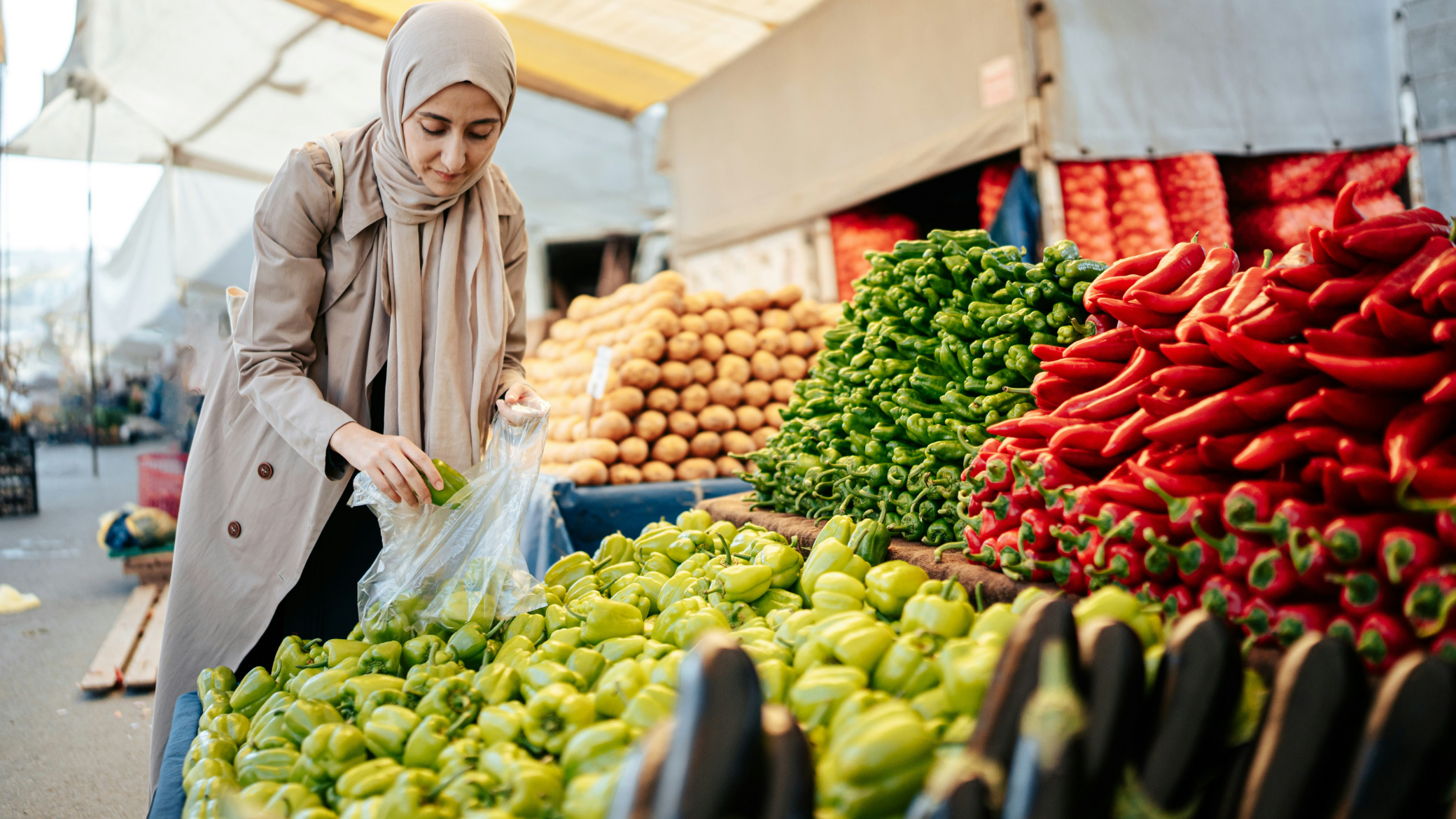 A woman wearing a beige coat and hijab shopping for green bell peppers at an outdoor farmer's market, with stacks of red and green peppers and other vegetables displayed on tables.