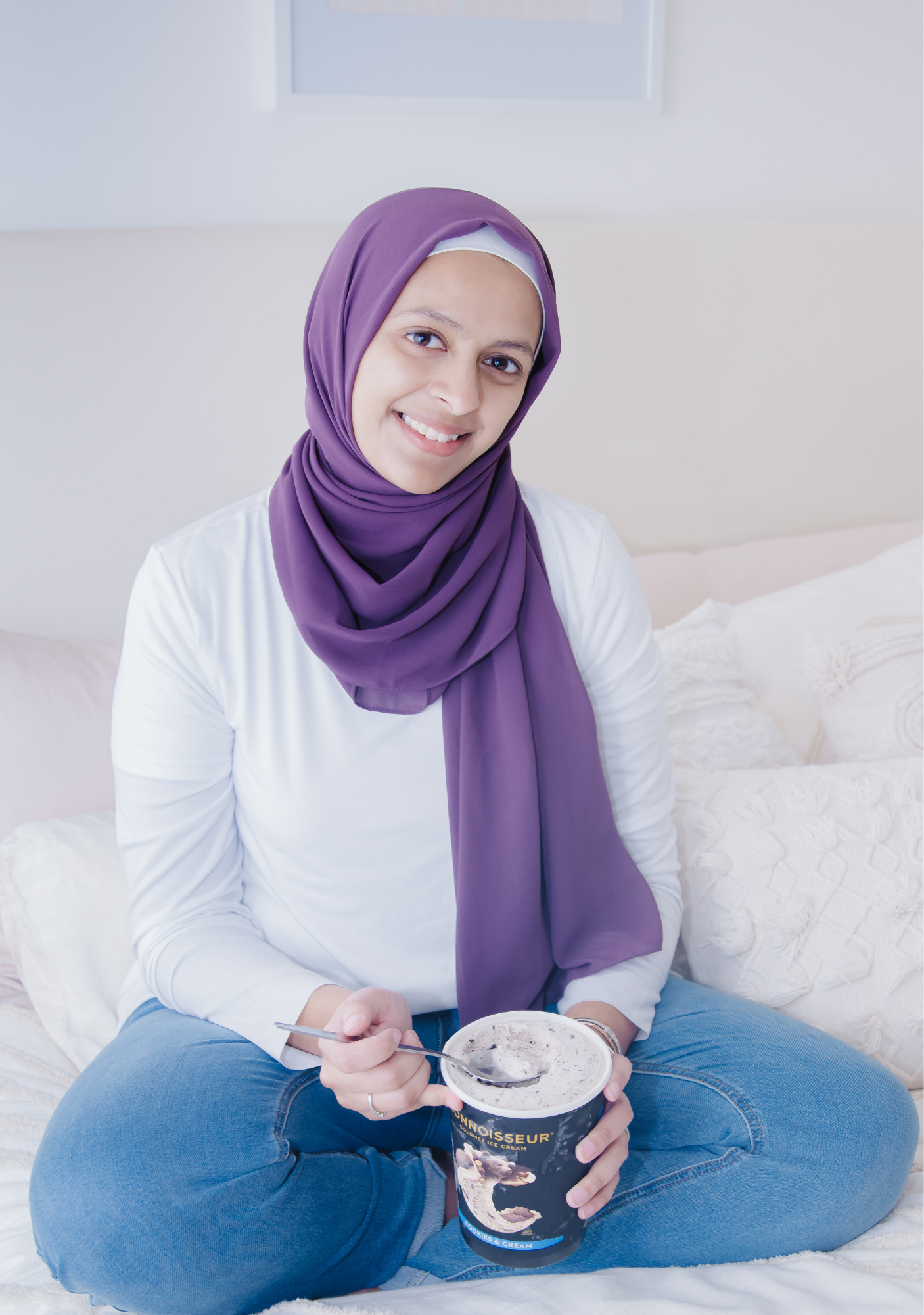 A young woman wearing a purple hijab and white shirt is sitting cross-legged on a bed, holding a cup of ice cream and smiling at the camera.