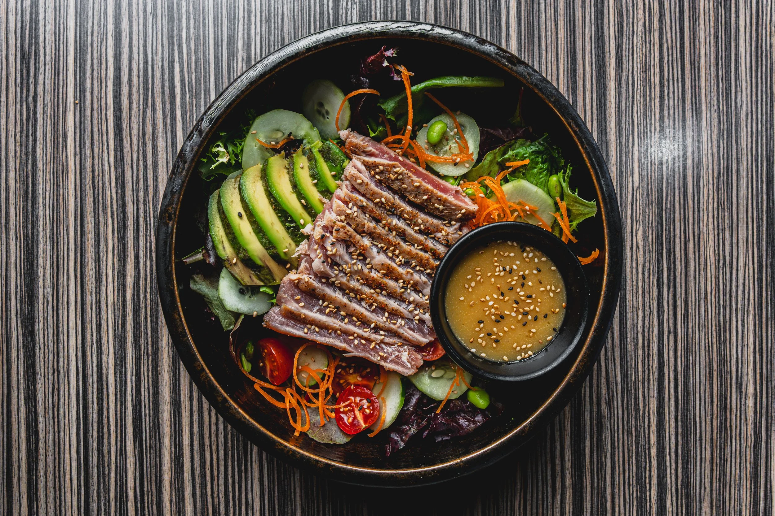 A poke bowl with sliced avocado, seared tuna, cherry tomatoes, cucumber, shredded carrots, mixed greens, and a side of sesame seed dressing.