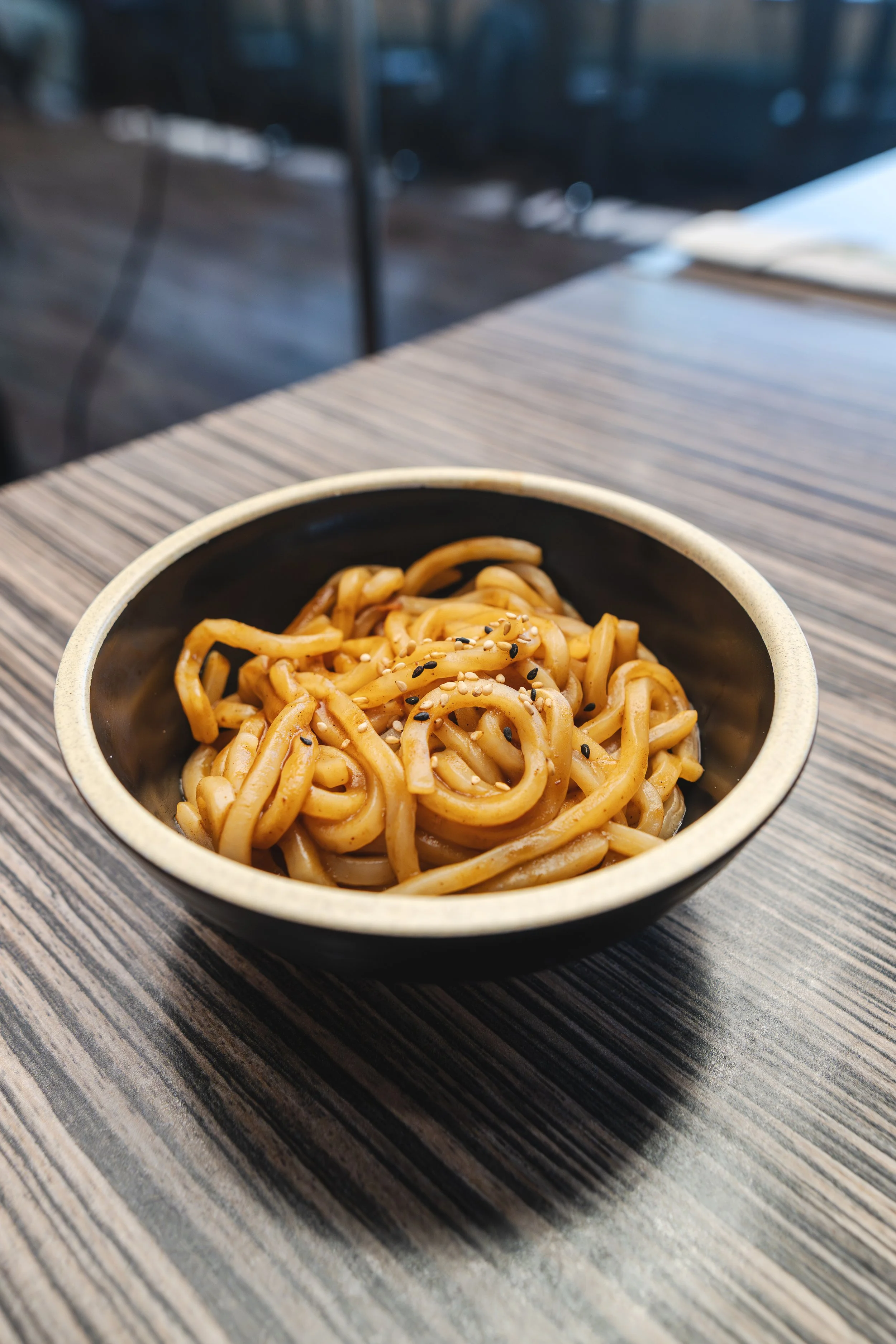 A bowl of stir-fried noodles topped with sesame seeds on a wooden table.