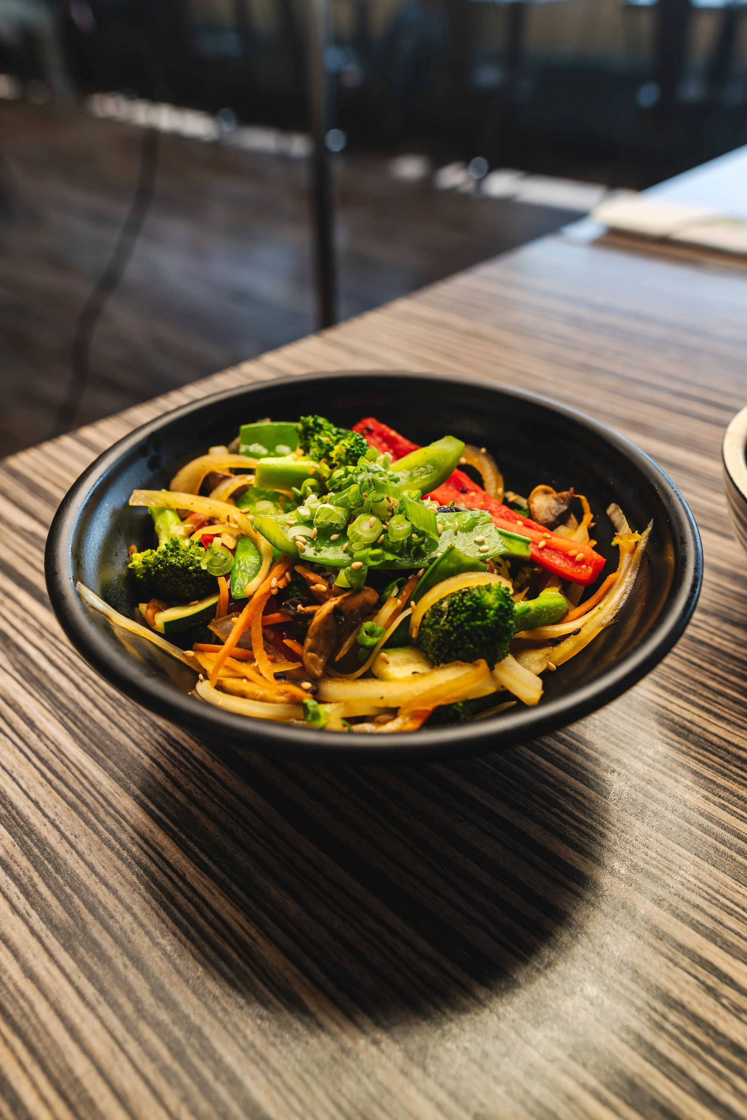 A black bowl of colorful stir-fried vegetables including broccoli, red bell peppers, snap peas, carrots, and mushrooms on a wooden table.