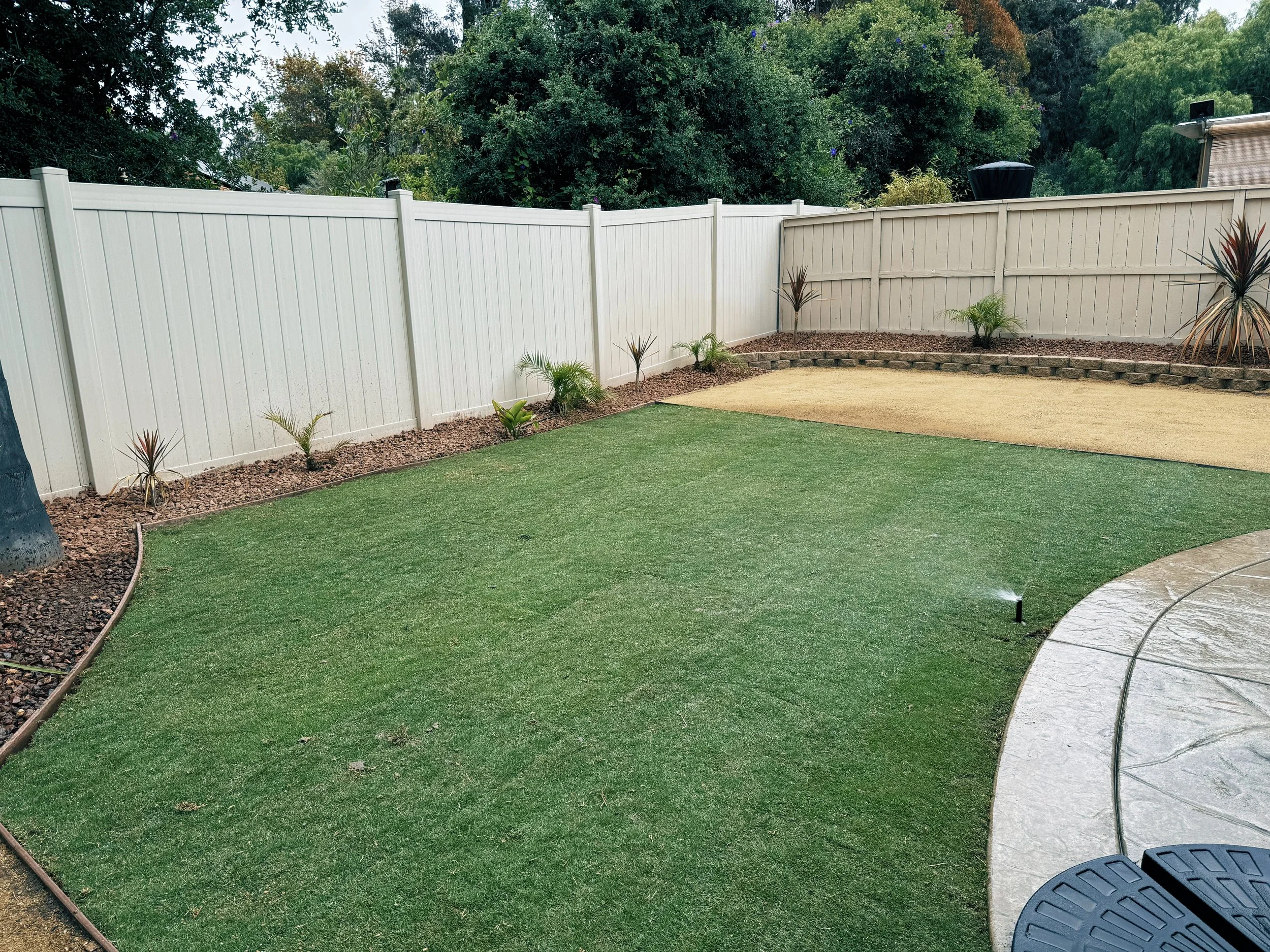 Backyard with artificial grass lawn, decorative plants, and a white vinyl fence with a garden bed.