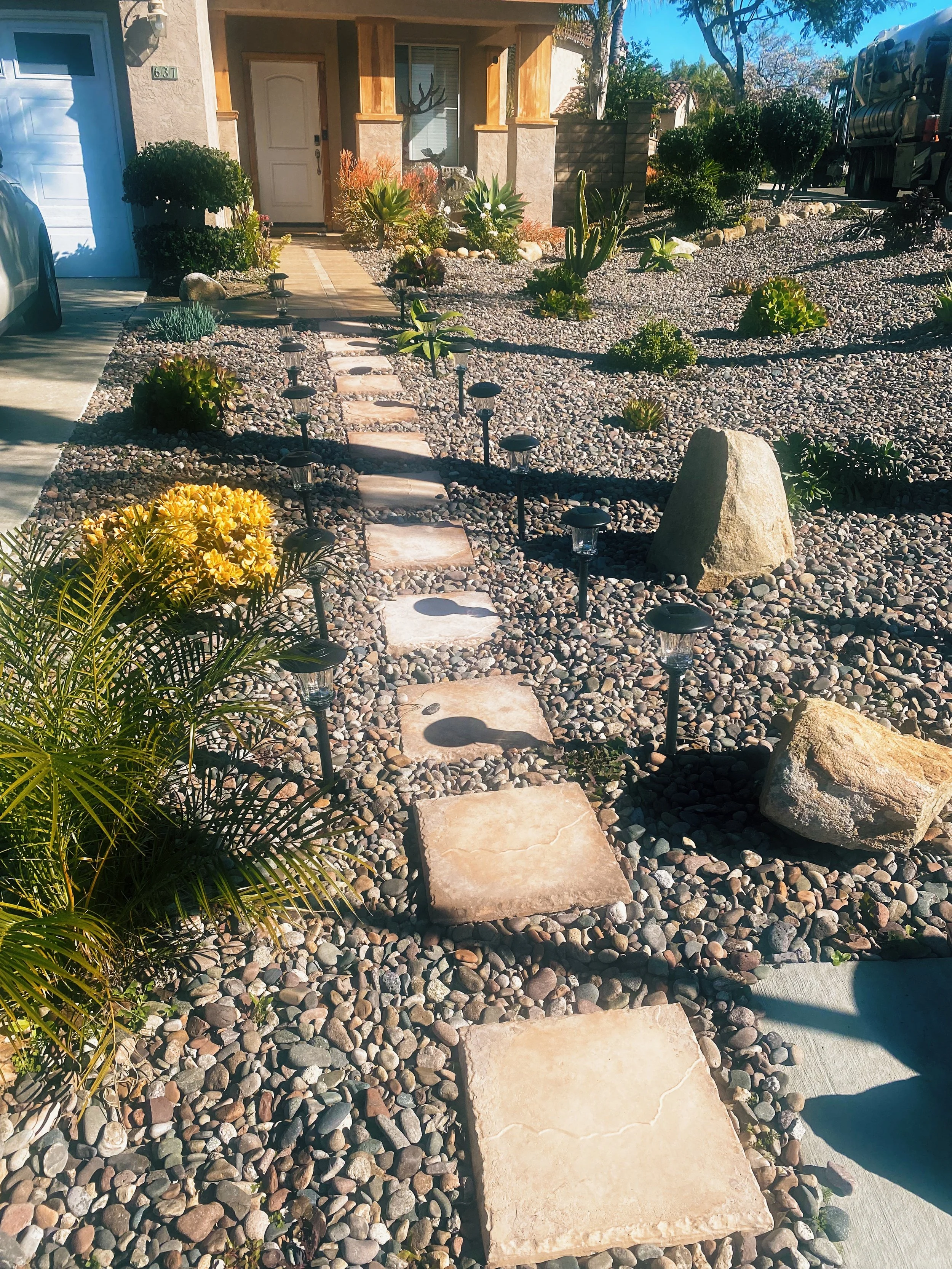 Front yard with a stone pathway lined with small solar-powered lights, various desert plants, and large rocks, leading up to a house with a porch. The house has a white door, a garage with a blue door, and a welcome sign. A car is partially visible on the driveway, and there is a large truck parked on the street.